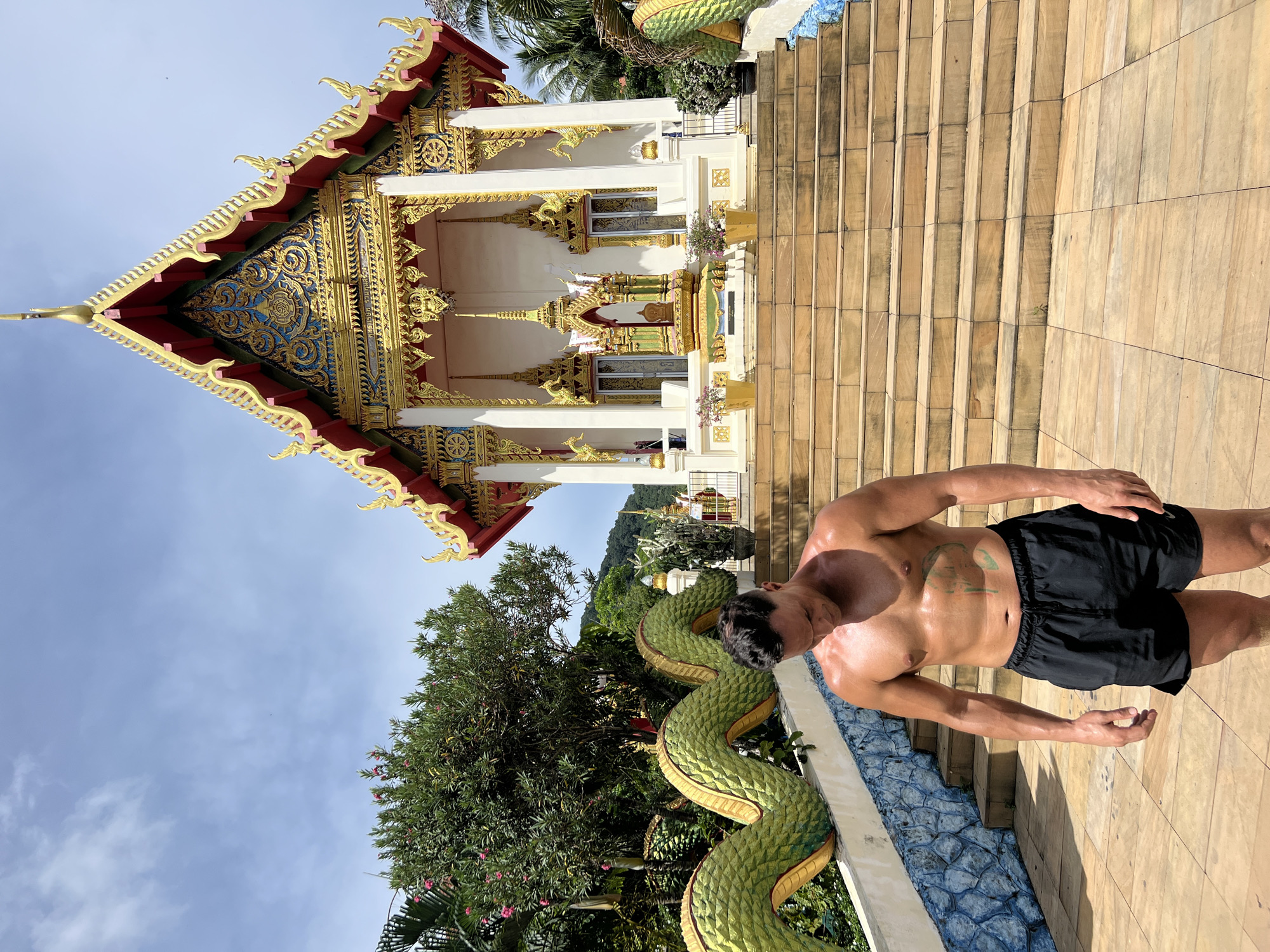 The image shows a shirtless man standing on a wooden staircase outside a temple in Karon, Thailand. The man is tanned and appears to be sweating, with a tattoo visible on his chest. He is wearing black shorts. To his left, a green, scaly serpent sculpture with yellow accents winds its way up the stairs. The temple itself is ornate, with a golden roof and intricate carvings. Palm trees and lush green foliage are visible in the background. The sky is a clear blue with a few white clouds, suggesting it is daytime and the weather is warm. The overall scene conveys a sense of tropical warmth and cultural richness. There is no visible text in the image.
