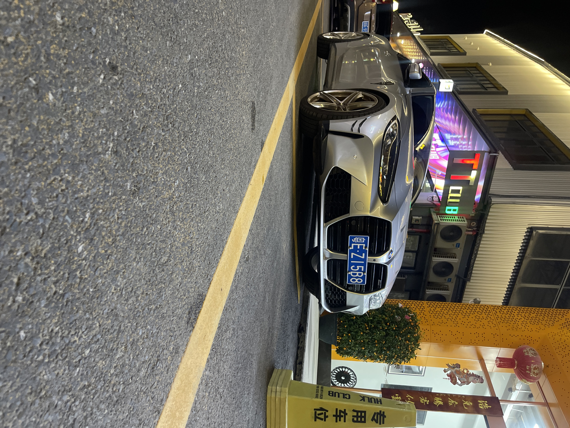 This media file captures a nighttime scene on a street, likely in Panyu District, China. The primary subject is a sleek, silver BMW parked alongside the road. The car features distinctive front-end styling, a license plate reading "粤E-Z15B8," and custom rims.
The setting appears to be a commercial or entertainment area, as evidenced by the brightly lit building in the background adorned with neon signs, including one that reads "TT CLUB." The building's facade is modern, with horizontal paneling and large windows. Various air conditioning units are visible mounted on the exterior.
In the foreground, a yellow line demarcates the parking area on the asphalt road. Signs with Chinese characters are also visible, one of which appears to be a parking designation ("专用车位") and another mentioning "HULK CLUB." A small, bushy plant with orange fruits, possibly kumquats, is situated next to the car. A traditional red lantern hangs from the building, adding a cultural touch. The overall atmosphere is lively and urban, characteristic of a commercial street at night.