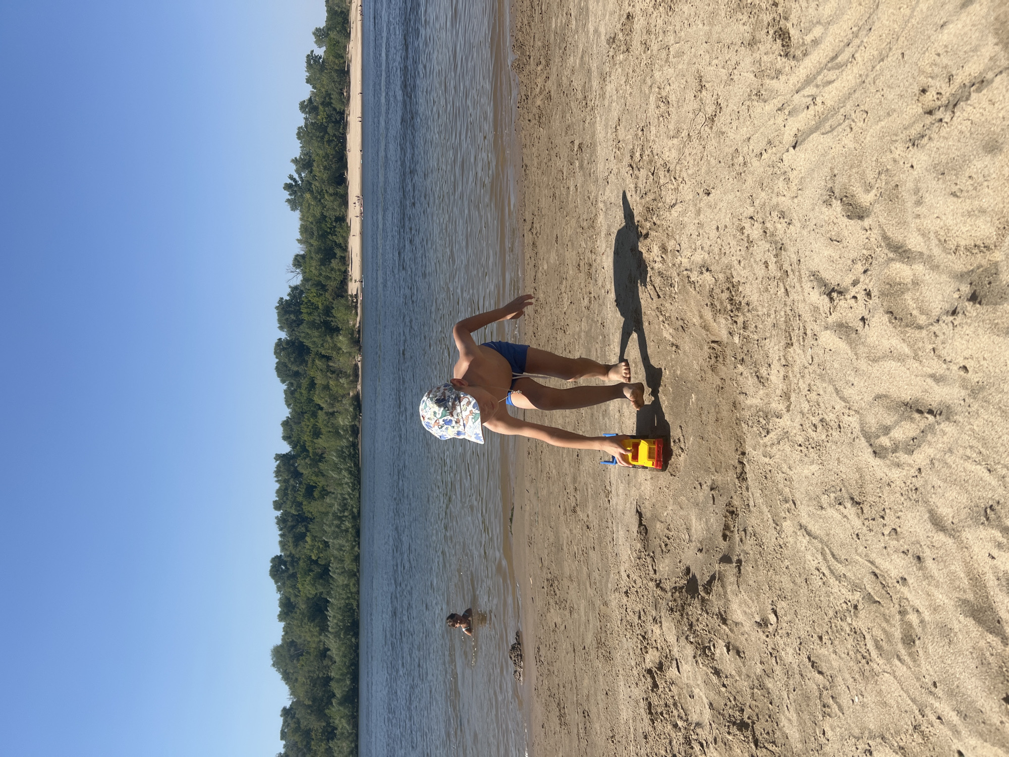 A young boy wearing a patterned cap and blue swim trunks is playing with a yellow and red toy truck on a sandy beach. He is in the foreground, bent over the truck. In the background, a calm river is visible, with trees lining the opposite bank. Another child is partially submerged in the water to the left of the boy. Several distant figures are scattered along the far shore, suggesting other beachgoers. The sky is clear and blue. The scene appears to be a natural river beach setting, possibly in or near Kilyakovka, Russia, given the presence of a river and wooded shoreline.