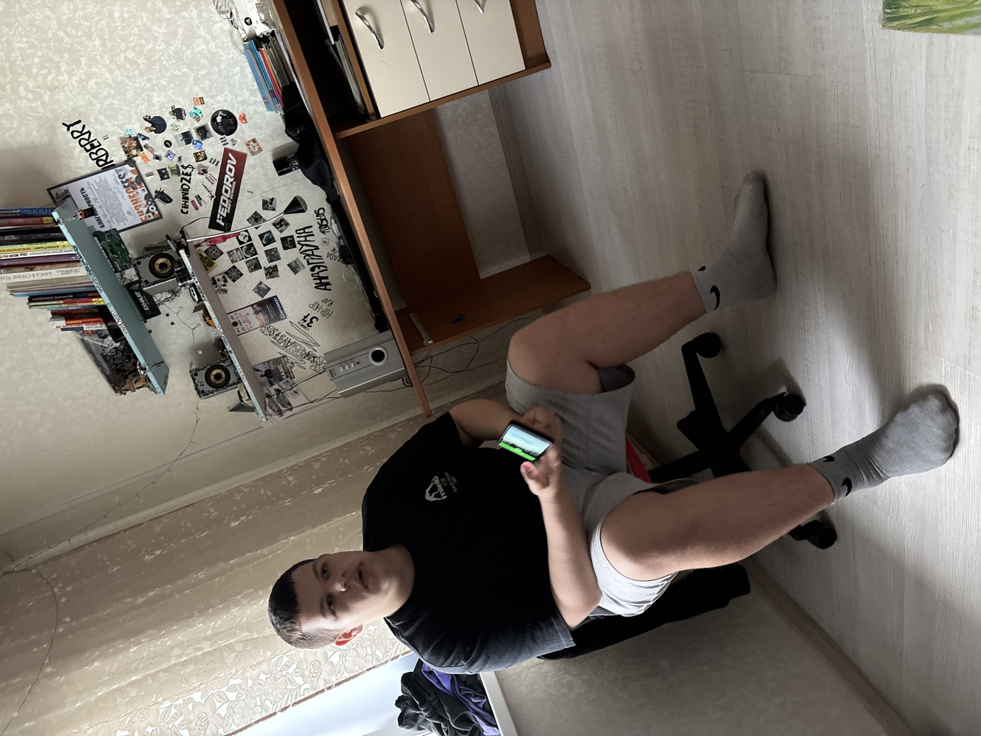 A young man is sitting in a chair and holding a smartphone. He is wearing a black t-shirt and grey shorts and grey socks. The background shows a wall with shelves filled with books and various items. A wooden desk with drawers is also visible, along with a computer monitor and speakers. The room appears to be a study or bedroom.
