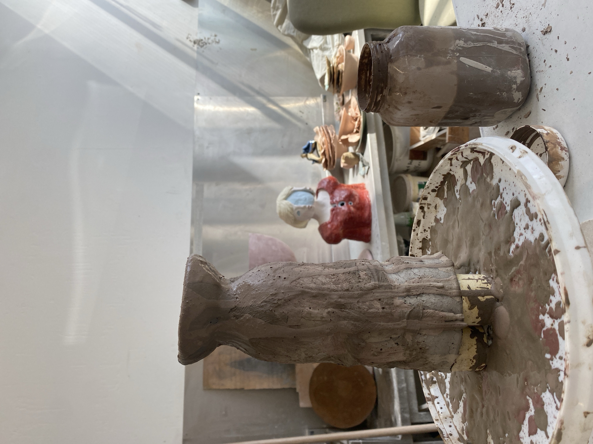 The image shows a ceramic studio, possibly in Saint-Ouen-sur-Seine, France. A clay sculpture on a dirty white wheel is the primary subject. The sculpture appears unfinished, with a roughly shaped body and head. Behind it, there is a painted clay bust of a woman, dressed in a red top and with light hair. Other clay figures and pottery are also visible in the background, including a dirty glass jar on a white surface. The surfaces in the room show signs of clay residue. The lighting is bright and appears to be natural. The background includes a metallic wall or shelf.