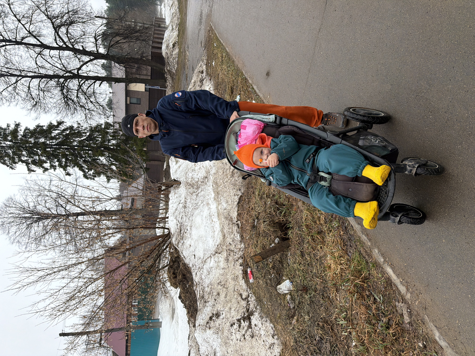 A man wearing a navy blue jacket, black beanie, and orange pants is standing behind a stroller on a paved path. The stroller contains a child wearing a bright orange hat, teal jumpsuit, and yellow rubber boots. The child has an orange lollipop in its mouth. The path runs alongside a dirty, melting snowbank, and the background shows houses and bare trees. The scene appears to be outdoors, possibly in a city like Chaikovsky, Russia.