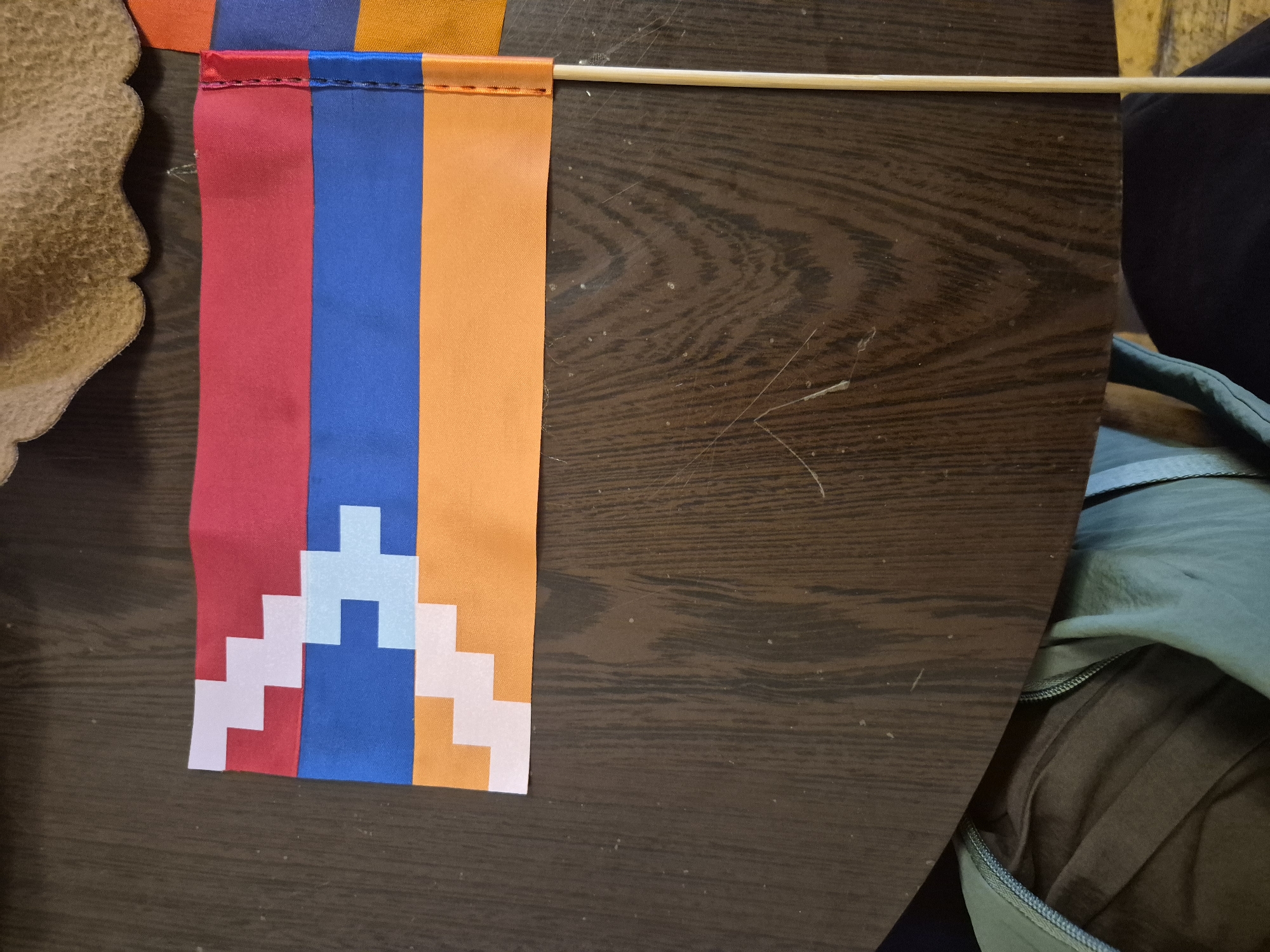 The image displays a close-up, overhead view of two small flags, one partially obscured by the other, resting on a dark wooden table. The prominent flag on the left features horizontal stripes of red, blue, and orange, with a white, stepped zigzag pattern superimposed on each stripe. The flag on the right, partially visible, appears to share a similar design. A thin wooden stick, likely a flag pole, extends vertically from the bottom right corner of the frame, attached to the edge of the prominent flag. The wooden table surface is visible throughout, showing a wood grain texture and some dust or debris scattered on it. The background is blurred, showing the edge of what might be a curved object, possibly a hat or a piece of fabric, in the upper right. The lower part of the image shows folded fabric, likely clothing, in shades of dark green and brown, hinting at a person sitting at the table. There is no discernible information about the city of Yerevan, Armenia, within the image itself.