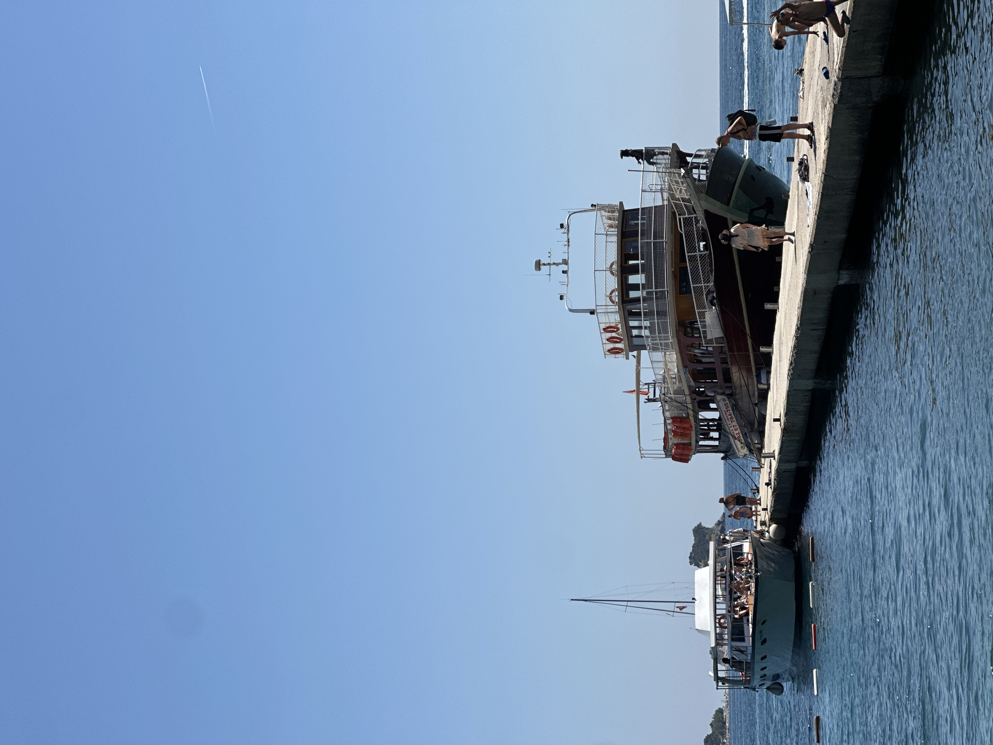 This image captures a sunny day at a pier in Budva, Montenegro, with two boats docked. A larger, older wooden boat with life preservers visible is moored to the right, while a smaller, white passenger boat is on the left, alongside a sailboat mast. Several people are on the pier, some standing and observing, while others appear to be disembarking or preparing to board. A few individuals are in the water, enjoying the blue sea under a clear sky with a faint contrail from an airplane. The pier itself is concrete and extends into the water, creating a bustling scene of maritime activity.
