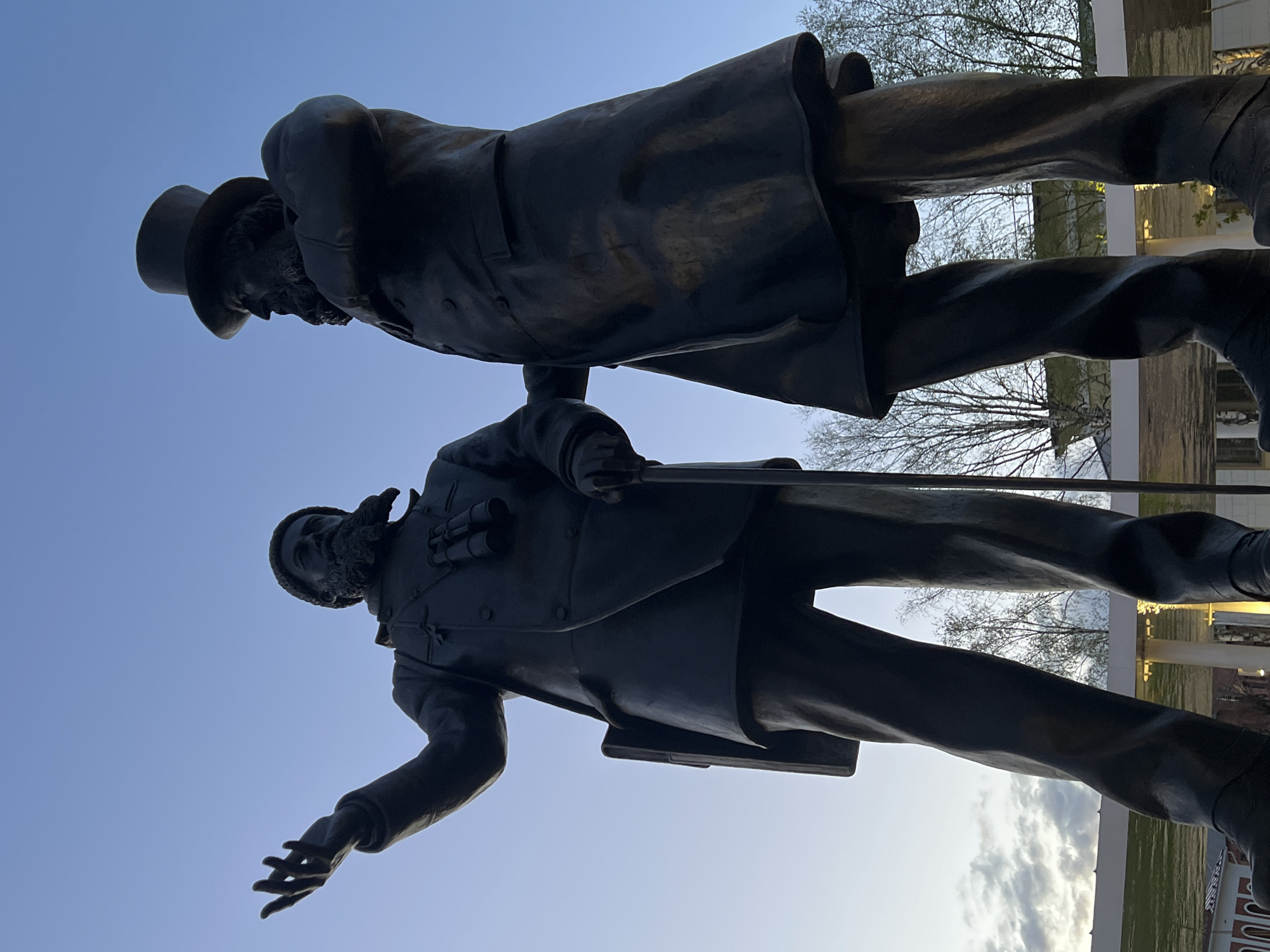 The image shows a bronze statue of two men, presumably historical figures. One man is wearing a top hat and a long coat. The other man is wearing a jacket and holding binoculars. They appear to be standing side-by-side. The background includes a blue sky with some clouds, bare trees, and parts of buildings.
Based on the pose and appearance of the figures, the statue may represent the meeting of two notable individuals, and given the location suggestion in Cherepovets, Russia, the figures are likely related to the history or development of that city. Additional details would be needed to definitively identify the specific figures.