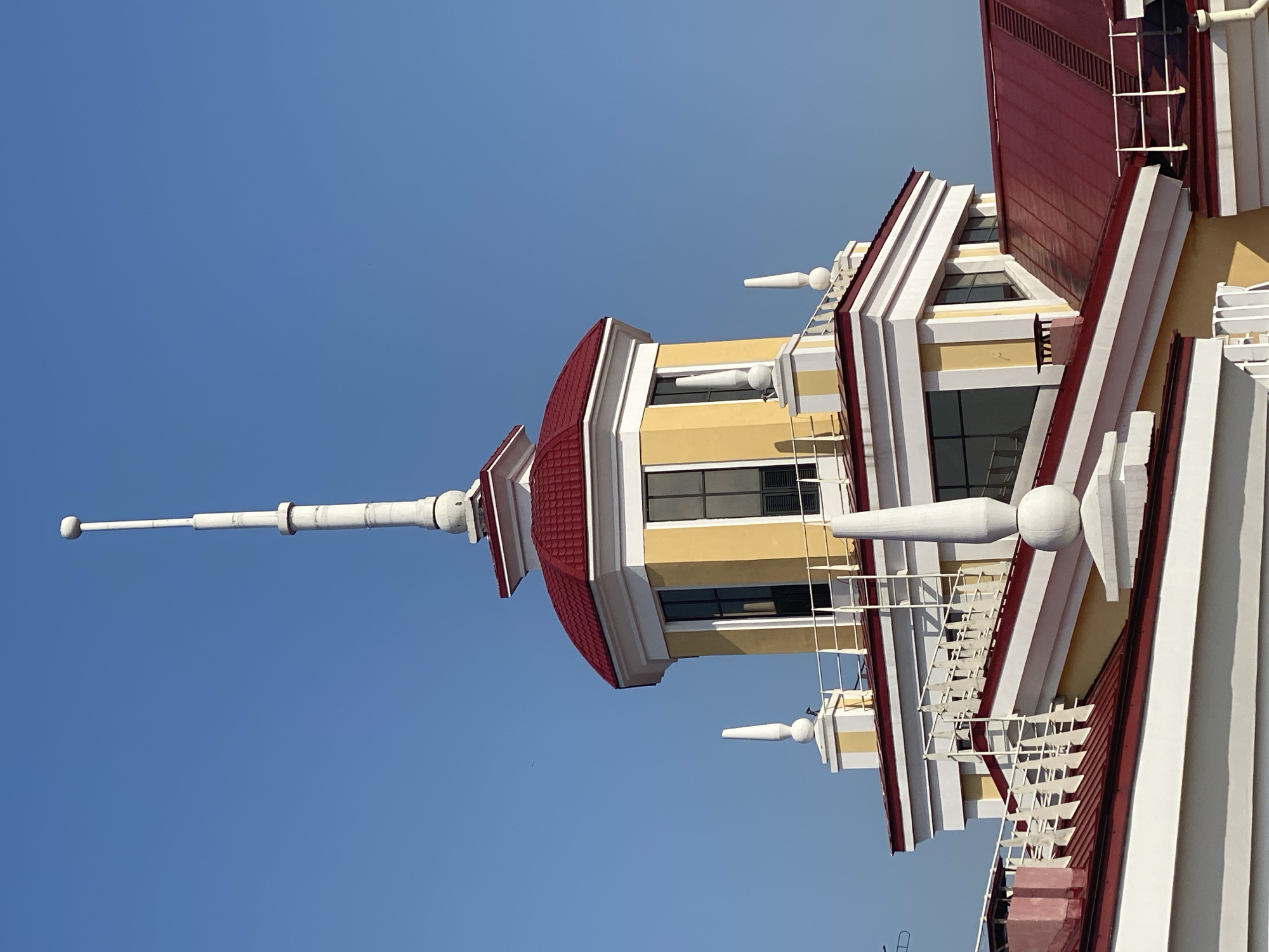 The image shows the upper portion of a building with a distinctive yellow facade and a red roof. The building features a central tower topped with a spire and a ball-shaped ornament. Several decorative white finials are also visible on the roof. The sky is clear and blue. The building appears to be part of a larger complex, with other sections of the red-tiled roof visible in the foreground. The overall architectural style suggests a formal or institutional building. The location is the Novokuznetsk Regional Drama Theatre in Novokuznetsk, Russia. There are no people visible in the image.