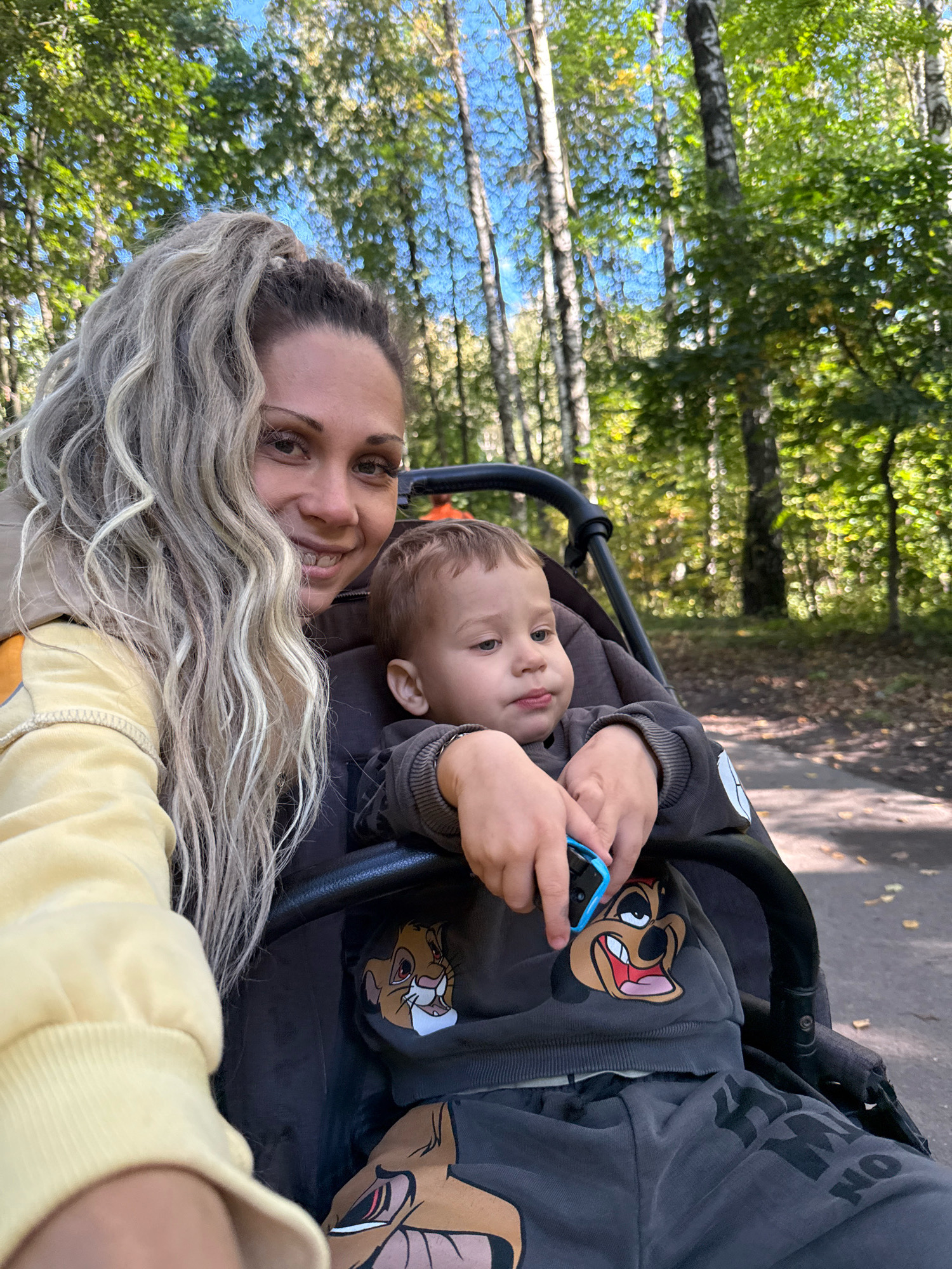 In an outdoor park setting, a woman with long, blonde, wavy hair tied up high smiles at the camera while taking a selfie. She is wearing a yellow hoodie with orange accents. Next to her, a young child is seated in a stroller, looking to the side. The child is wearing a dark gray sweatshirt with a graphic print of a cartoon character, likely from The Lion King, on the front and matching pants. The stroller has a black frame. The background consists of a path through a wooded area with trees, foliage, and sunlight filtering through the leaves. The overall scene depicts a casual outing in a park.
