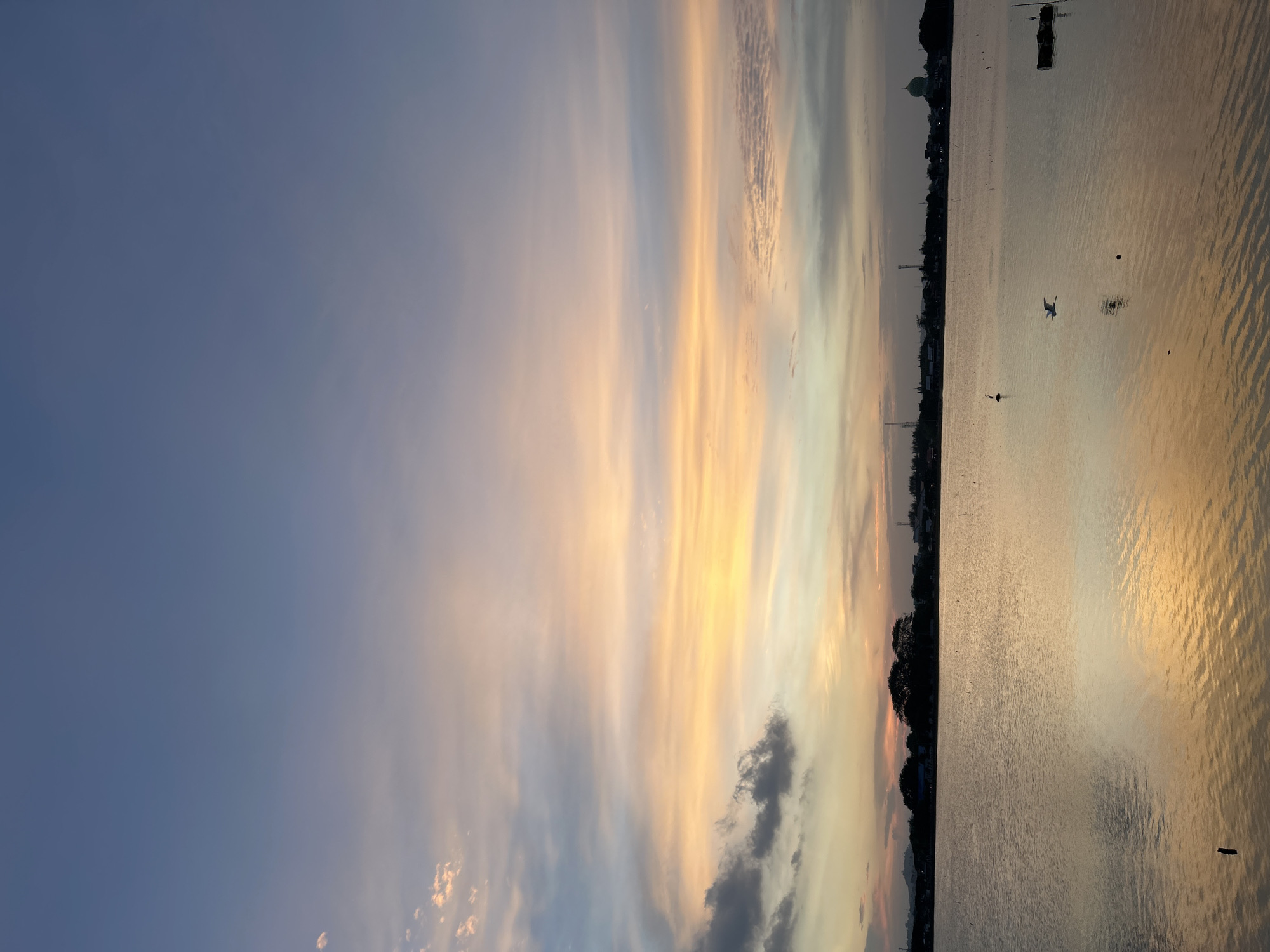 This image depicts a serene sunset over the waters of Lhokseumawe, Indonesia. The sky is a gradient of blues, purples, and oranges, with streaks of clouds catching the fading sunlight. The calm water reflects the colors of the sky, creating a warm and inviting atmosphere. In the distance, a silhouetted skyline of buildings, trees, and a mosque dome line the horizon. A few birds are visible in the water, with one in flight, adding a sense of life to the tranquil scene. The overall impression is one of peace and natural beauty at dusk in Lhokseumawe.