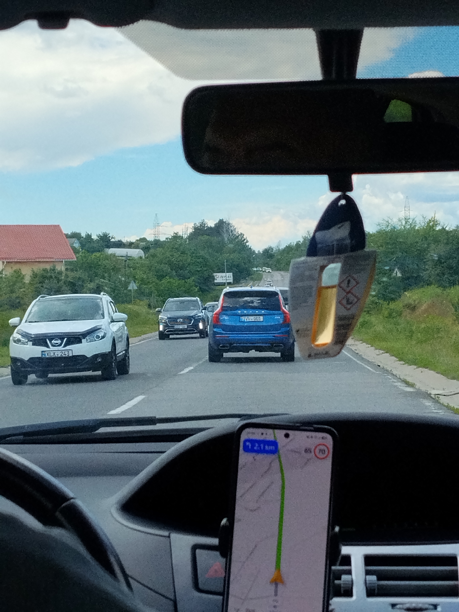 The image is taken from the interior of a vehicle, looking forward through the windshield. The scene depicts a highway in Budești, Moldova, on a cloudy day. Several cars are visible on the road, including a white Nissan Qashqai in the foreground with the license plate "XLX-243", a dark SUV and a blue Volvo SUV further ahead. A car air freshener hangs from the rearview mirror. A smartphone displaying a navigation app is mounted on the dashboard. The surrounding environment includes trees and power lines.