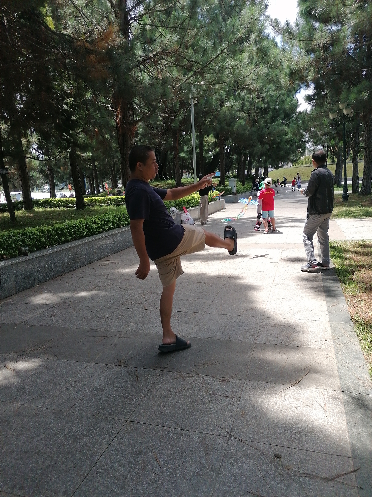 A man in a dark blue t-shirt and tan shorts is captured mid-action in a park setting. He is balancing on his left leg, with his right leg extended outwards, as if performing a kick or a dance move. His left arm is outstretched to his side, and his right arm is bent at the elbow, with his hand near his chest. The man is wearing dark colored sandals on both feet.
Behind him, further into the park, several other individuals are visible. To the right, a man in a dark jacket and light-colored pants is standing and holding a white object, possibly a kite string. In the middle ground, a young person in a red t-shirt and blue shorts, wearing roller skates, is holding onto a kite that is partially visible. Several other figures, including children and adults, are scattered throughout the background, suggesting a public gathering or recreational area.
The environment appears to be a well-maintained park with lush green trees lining a paved walkway. The walkway is made of large, grey stone tiles. A low, grey stone wall with a manicured hedge borders the left side of the walkway. Sunlight filters through the trees, creating dappled shadows on the ground.
The scene is likely located in Da Lat, Vietnam, a city known for its pine forests and numerous parks, popular for outdoor activities and recreation. The overall atmosphere is one of leisure and outdoor enjoyment.