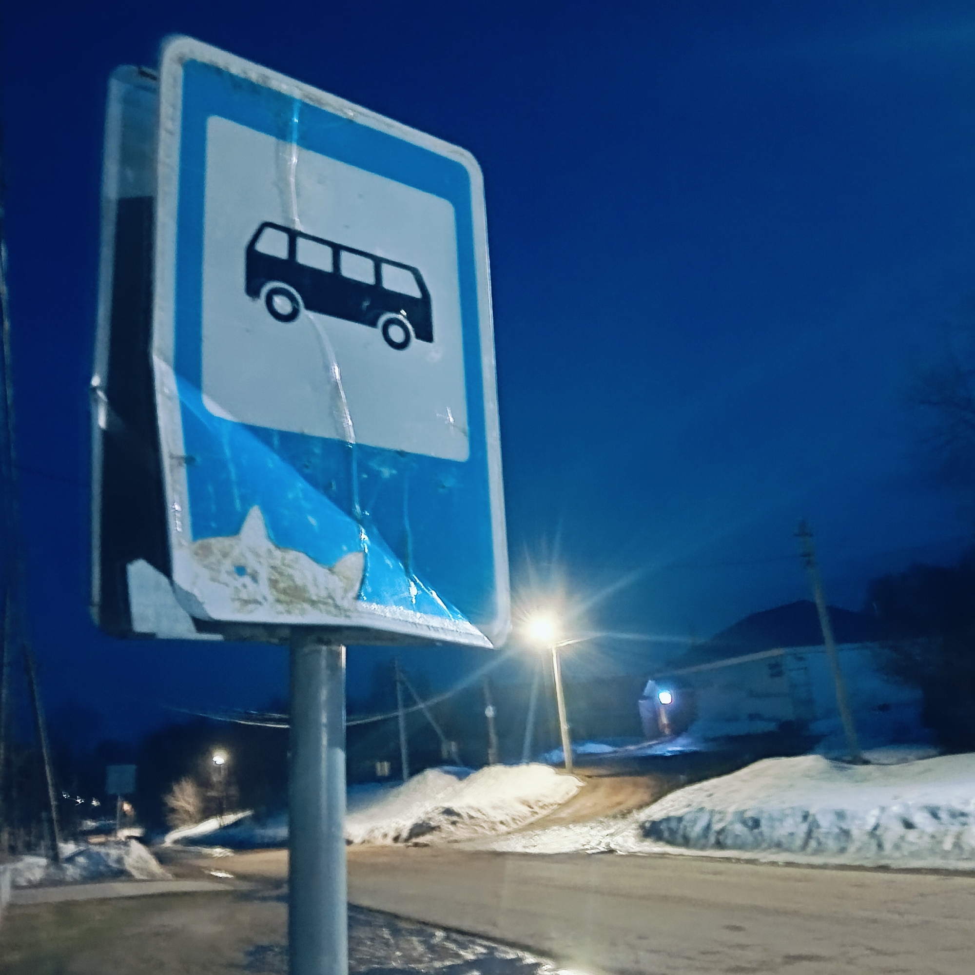The image shows a bus stop sign located in Зипуново, Russia. The sign is blue and white, depicting a black bus. The sign is weathered and peeling, and is mounted on a grey metal pole. The surrounding environment includes a road, snow-covered ground, a street lamp, and a building in the background. The time of day appears to be dusk or night.