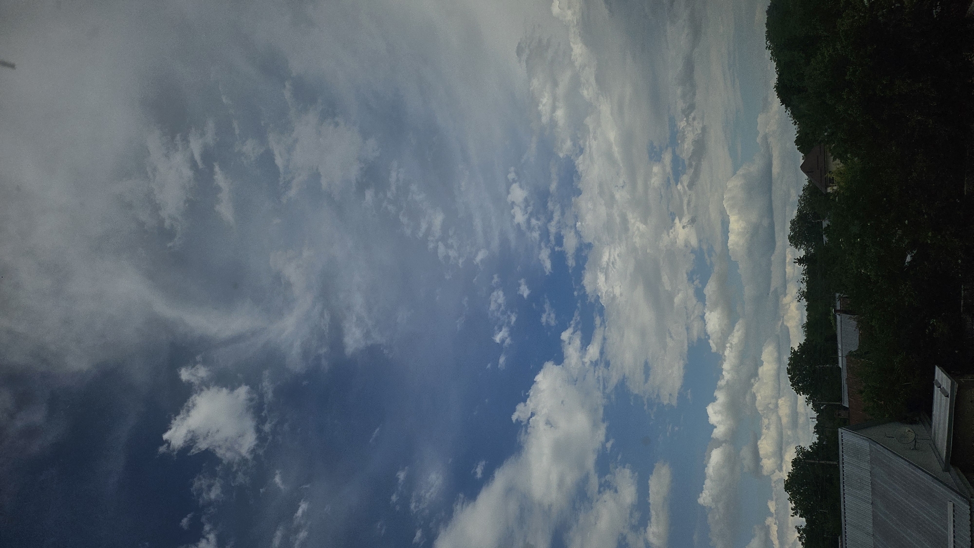 The image depicts a view of the sky above a residential area, likely in a suburban or rural setting, given the presence of trees and rooftops. The sky is mostly filled with clouds of various densities, ranging from scattered white cumulus clouds to larger, more diffused grey formations. The lighting suggests it is daytime, possibly late afternoon or early morning, as the light is somewhat diffused.
In the foreground, parts of several rooftops are visible. On the left, a metal-sheeted roof with a peaked structure dominates the lower left corner. To its right, another rooftop with darker tiles is partially obscured by trees. Further to the right and in the background, more rooftops and a portion of a building with a distinctive brown, conical roof can be seen peeking through the dense green foliage of trees. The trees form a thick canopy across the middle and lower portions of the image, suggesting a well-treed environment.
There are no people or obvious signs of recent activity. The scene is static and captures a moment in time. The overall impression is one of a quiet, overcast day in a vegetated area.