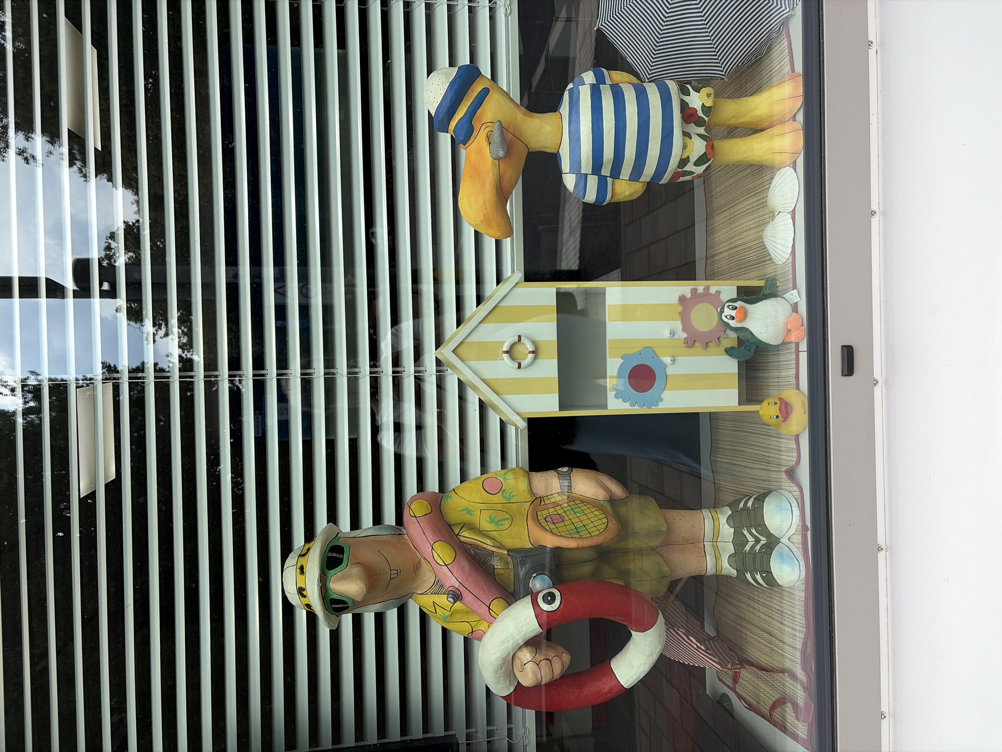 The image shows a shop window display in Würzburg, Germany, featuring beach-themed figures. On the left is a large, carved figure of a man wearing a safari hat, sunglasses, a yellow shirt with palm trees, and khaki shorts. He is holding a red and white life preserver ring and has a camera around his neck and a tennis racket in his hand. In the center of the display is a miniature yellow and white striped beach hut with decorations. To the right of the beach hut is a large, carved yellow bird figure wearing a blue and white striped shirt, a sailor's hat, and colorful shorts. In front of the figures are a small yellow rubber duck, a plush penguin, and several seashells. An umbrella with blue and white stripes is visible to the right of the bird figure. The display is set against a backdrop of horizontal white blinds, with reflections of trees and sky visible through the window.