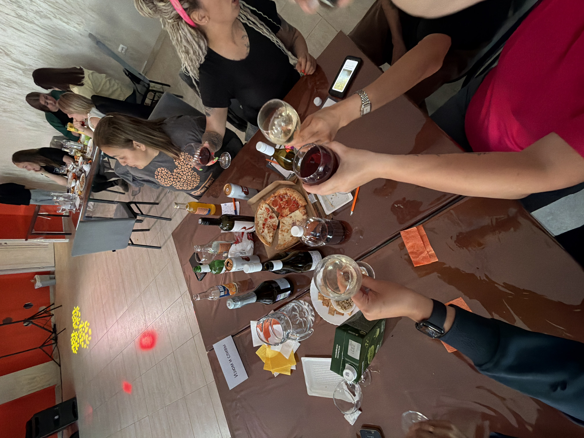 Here is a forensic description of the image:
**Location:** Indoor space, likely a private room or dining area in Chaikovsky, Russia. There are light reflections on the floor, suggesting a party or gathering with potential lighting effects.
**People:** The image captures several individuals. In the foreground, multiple people are seated around a table. There is a person with blonde dreadlocks wearing a black shirt. To their right, another person is wearing a bright pink shirt. Further down the table, a person is wearing a gray shirt with orange heart or leopard print. At the end of the table, there is a group of people in various colored tops.
**Objects:** A long table covered in a brown cloth dominates the center. The table contains a variety of items, including:
* Wine bottles, both empty and full.
* Wine glasses, some held aloft as if in a toast.
* A partly eaten pizza.
* A container of juice,
* Snacks on a plate.
* A sign with Cyrillic text "Изюм и Сымвы".
* Several smartphones
**Actions:** People are actively engaged in socializing and dining. The raising of wine glasses indicates a toast or celebratory moment. Consumption of pizza and drinking of wine are in progress.
**Interactions:** Close proximity and shared beverages indicate a social gathering. Toasts can be considered an interaction with the other people at the table.
**Environment:** The room appears to have tiled flooring with some colored lighting and red lights visible. Chairs are arranged around the table. Orange accents can be seen on the wall near what may be a speaker.