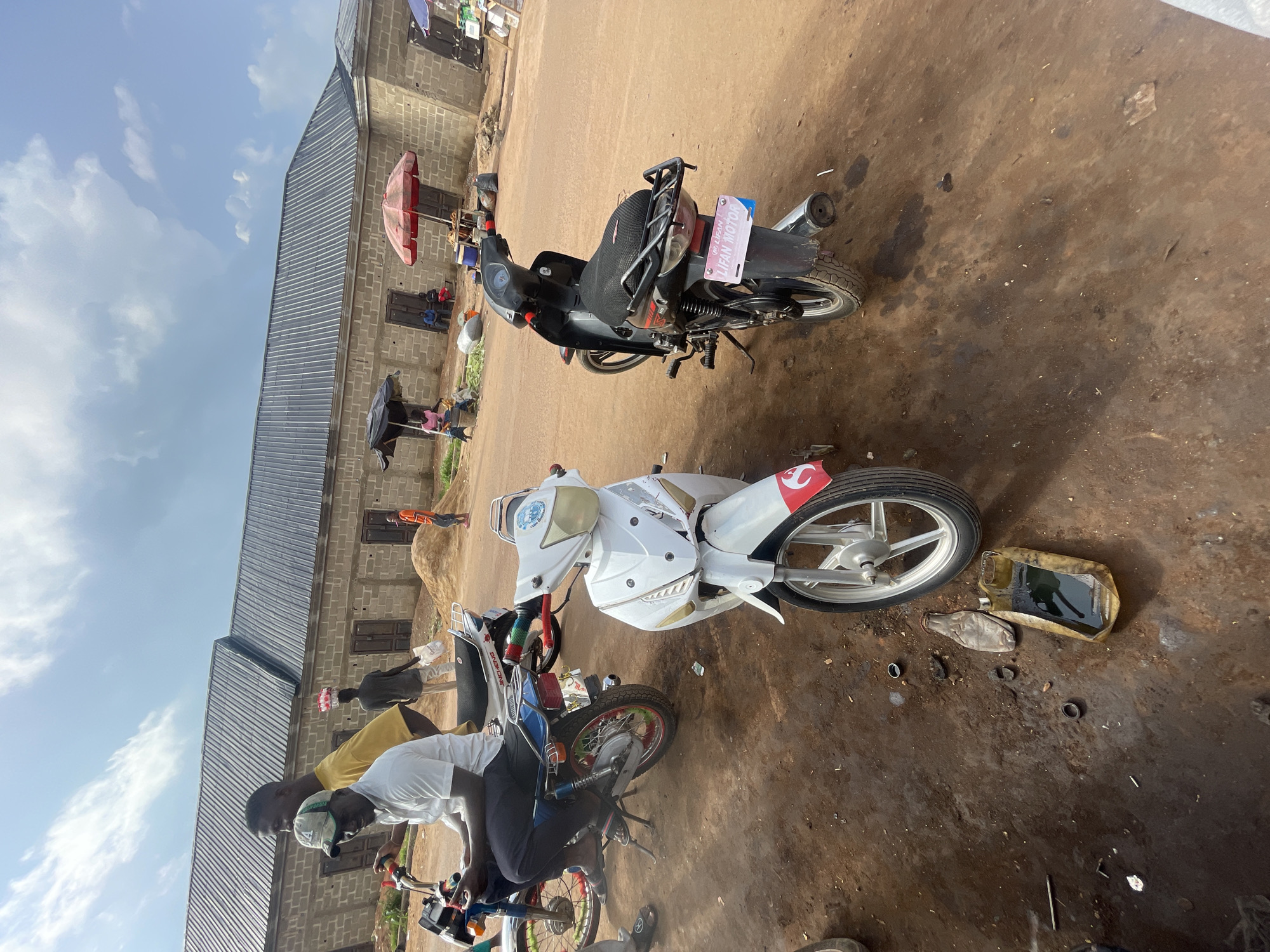 Two motorcycles are parked on a dirt ground in front of a building with a metal roof. One motorcycle is black with a pink license plate that reads "LIFAN MOTOR". The other motorcycle is white with red accents. There are a few people present, some standing and some sitting on the motorcycles. A red umbrella is set up near the building. The scene appears to be an outdoor market or workshop. The time of day appears to be during daylight hours. The weather seems to be clear with some clouds in the sky.