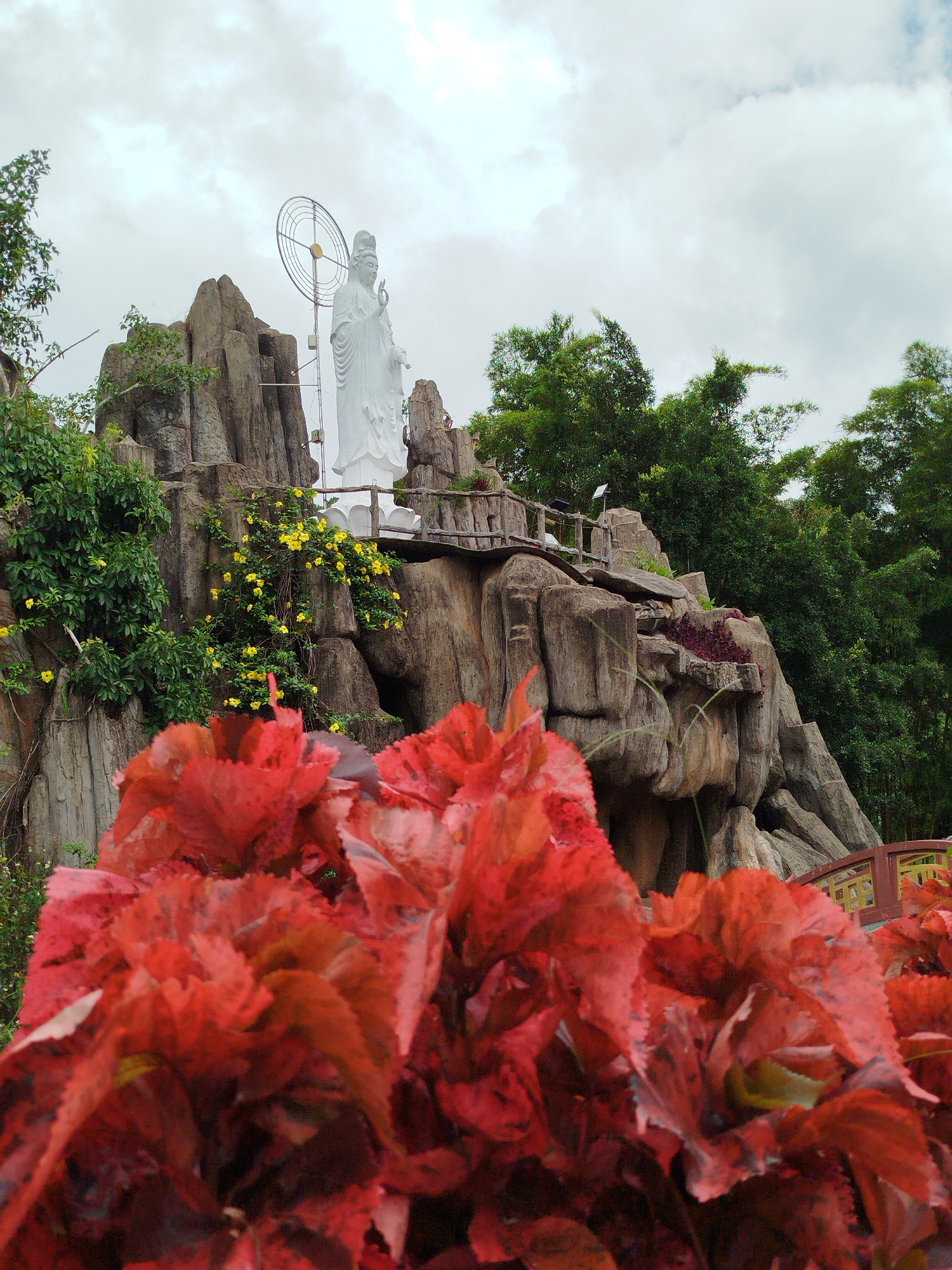 A full shot captures a white statue of a serene figure with a halo behind it, standing atop a rocky, tiered landscape in Xuan Huong - Da Lat, Vietnam. The statue is positioned centrally, overlooking a foreground filled with vibrant, out-of-focus red foliage. Behind the statue, a wooden bridge with a railing stretches across the rocks, and lush green trees form a dense backdrop against a cloudy sky. The rocky formations are textured and layered, with some areas adorned with small yellow flowers. The overall scene suggests a peaceful, elevated viewpoint within a garden or temple setting.