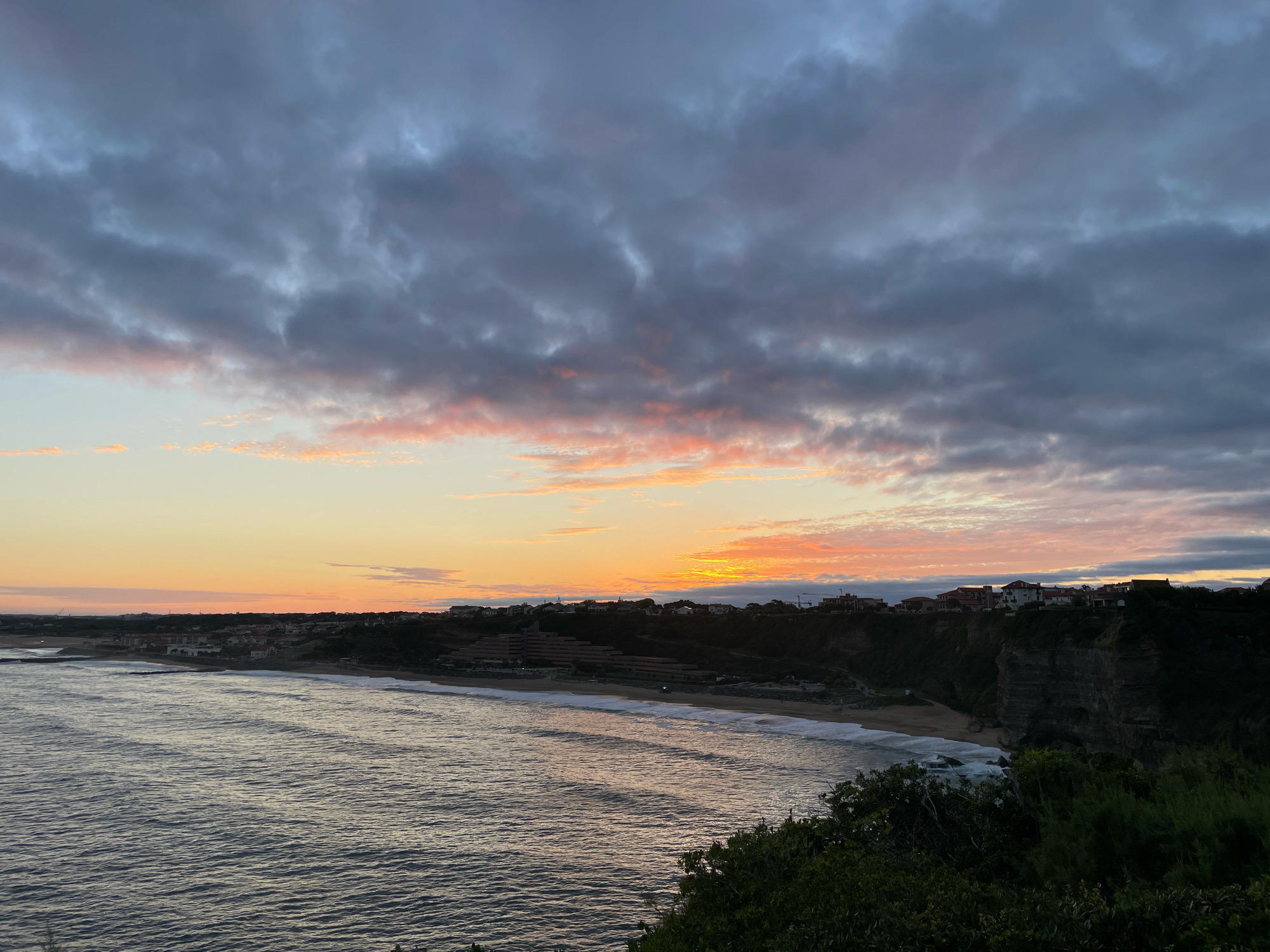 A wide shot captures a coastal view at dusk in Biarritz, France. The sky is filled with dramatic, dark gray clouds interspersed with streaks of pink and orange from the setting sun. Below the sky, a calm ocean stretches out, with gentle waves creating ripples and white foam along the shoreline. A sandy beach borders the water, backed by a cliffside dotted with buildings. These structures appear to be a mix of residential homes and larger buildings, some with red-tiled roofs, nestled into the hillside and along the clifftop. The foreground is dominated by dark green foliage, suggesting the vantage point is from a higher elevation overlooking the scene. The overall impression is one of serene natural beauty mixed with a hint of the urban landscape.