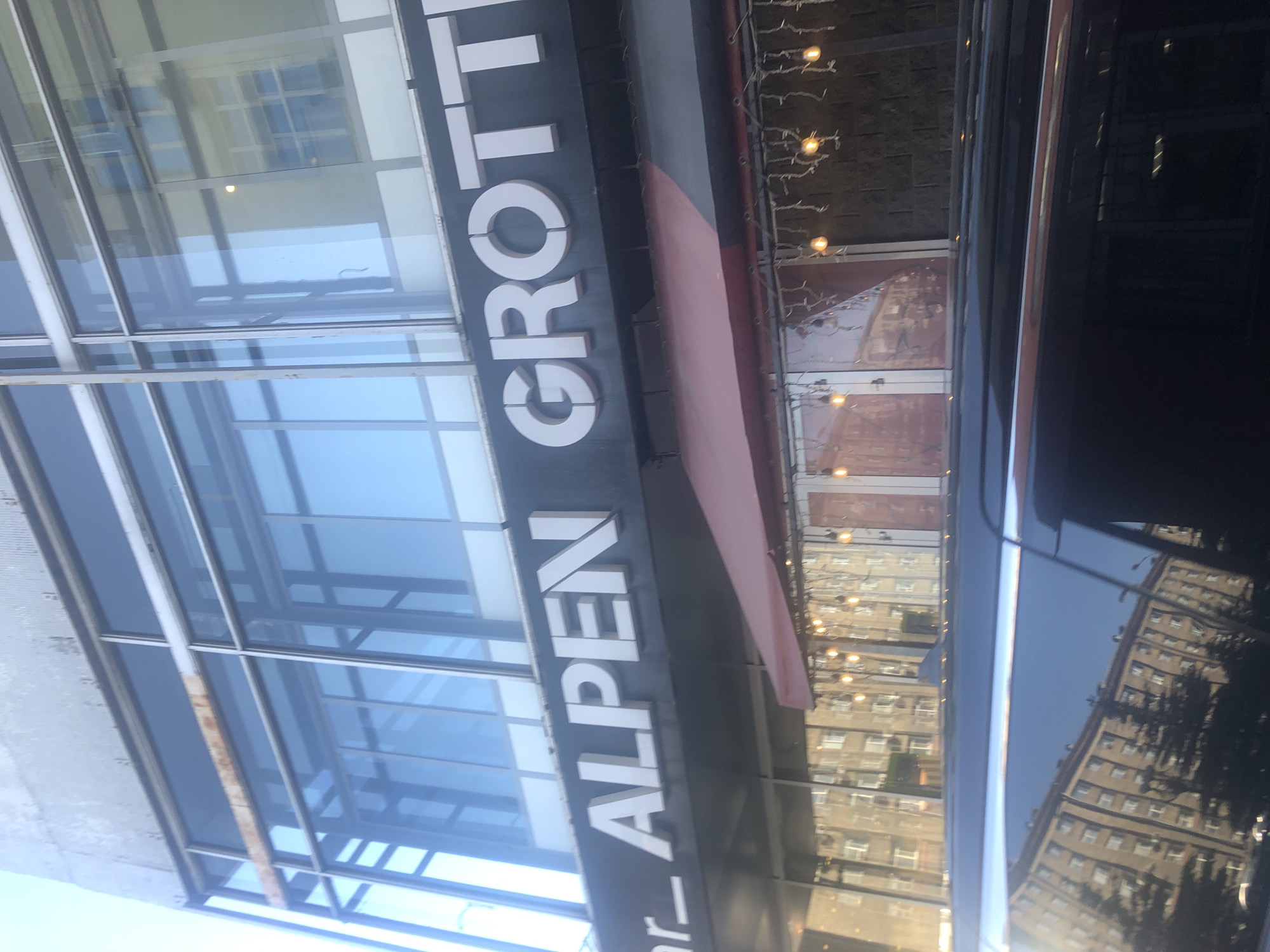 A sign on the facade of a building reads "ALPEN GROTTI". The sign is white lettering on a dark background. The building has large glass windows. Below the sign, there is an awning with string lights hanging from it. A black car is parked in front of the building, and the reflection of buildings and trees can be seen in its windows. The sky is visible at the top of the image.