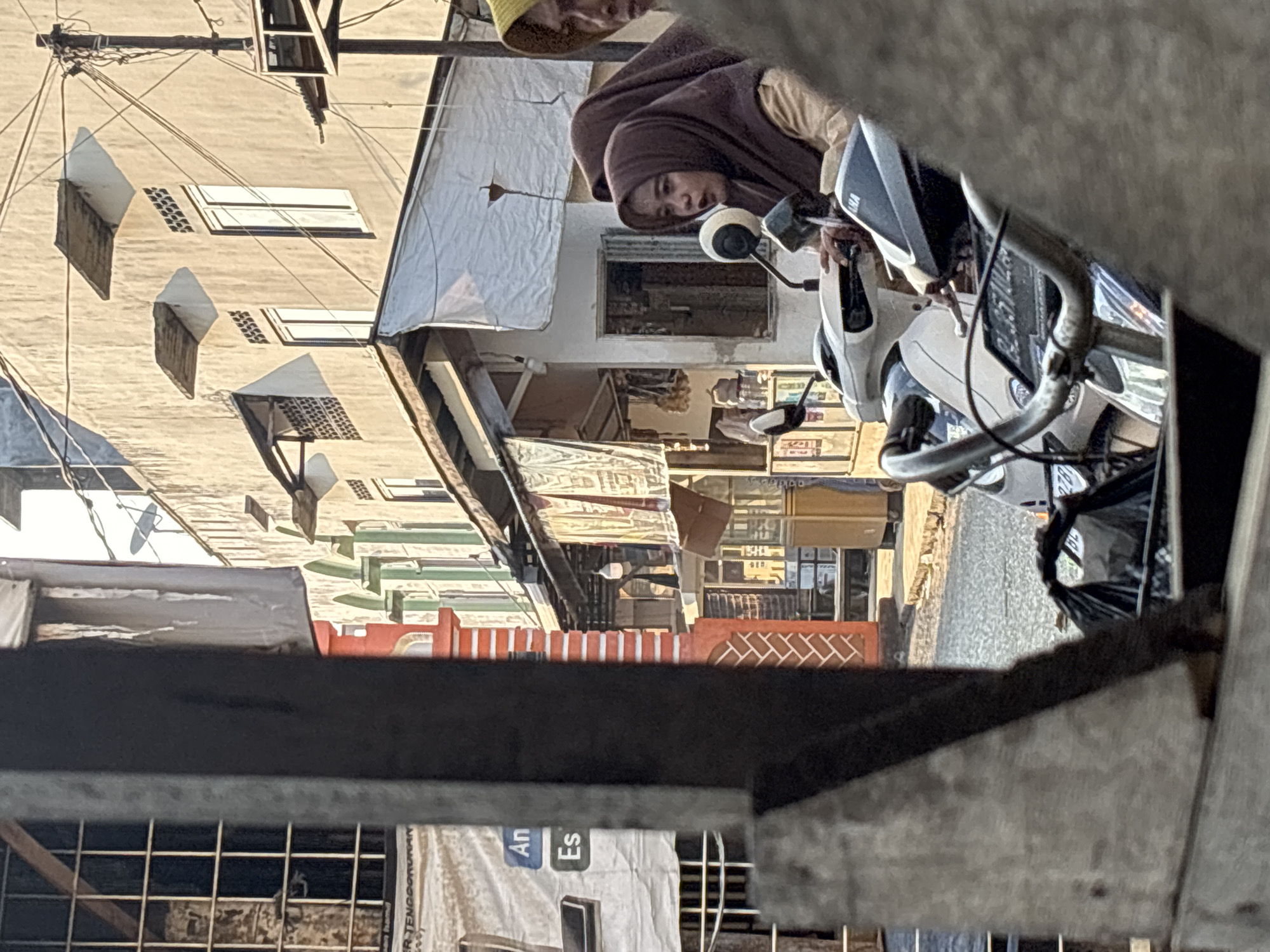 The image captures a vibrant street scene in Sangso, Indonesia, viewed from an obscured, slightly elevated perspective, possibly through a window or gate opening. The primary subject is a person seated on a white scooter, positioned towards the right of the frame. This individual wears a brown head covering, likely a hijab, and a light-colored top, looking directly at the camera with a neutral expression. Partially visible behind them is another person wearing a yellow hat.
The scooter is white with black trim, and a blurry license plate is visible on its front, displaying some unreadable characters and numbers, possibly beginning with "B 3310". Faintly, "YAMAHA" can be seen on the scooter's front panel.
In the background, a bustling street or alley stretches out, lined with multi-story buildings. The building on the left features a light-colored facade with several rectangular windows and protruding structures, possibly awnings or decorative elements, creating distinct shadows. Overhead, numerous utility wires crisscross the view. Further down the street, a series of colorful storefronts in red, green, and brown hues suggest shops or market stalls.
The scene is illuminated by bright daylight, casting clear shadows that indicate sunny weather, likely either morning or late afternoon. The overall atmosphere is that of a typical local street in a Southeast Asian town.
In the bottom left foreground, on what appears to be a crumpled banner or poster, the fragmented words "An" and "Es" are visible in a distinctive font, reminiscent of the "Breaking Bad" television series logo.