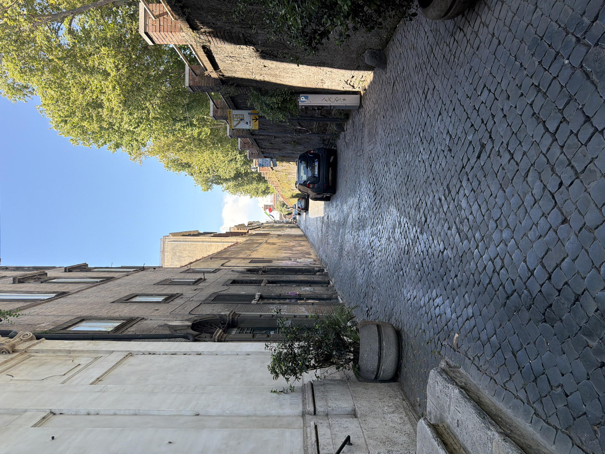 The image depicts a narrow, cobblestone street in a European city under a bright blue sky with scattered clouds. The street is flanked by old, brick and stone buildings on both sides. On the left, a tall, light-colored building features a decorative cornice and a large stone planter with a leafy green bush. A stone staircase is visible at the bottom left. On the right, a dark stone wall lined with trees runs alongside the street, with a parking meter and some signage visible. A dark-colored SUV is parked on the right side of the street, further down. In the distance, more buildings and trees are visible, suggesting a dense urban environment. The overall scene is one of an old European street, possibly in Italy, given the signage and architecture.
