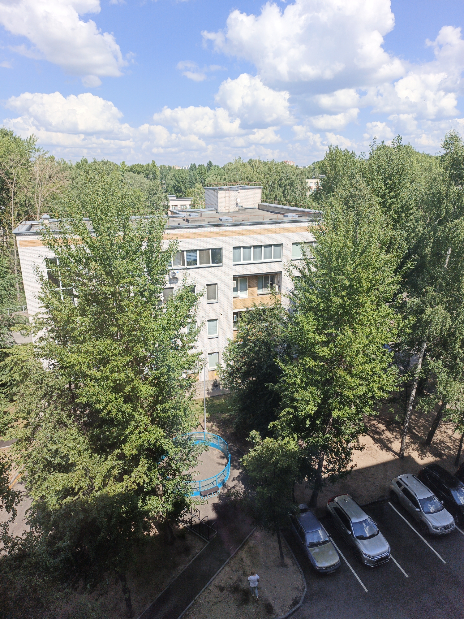This is an aerial, medium-range shot of a residential area in Kazan, Russia. The image features a multi-story brick apartment building with light-colored brick and orange trim, surrounded by dense green trees. In front of the building, there is a small, circular blue playground with a white border and benches. A parking lot is visible in the foreground with several cars parked, including a dark SUV, a silver SUV, a silver sedan, and a darker sedan. A person in a white shirt and blue pants is standing near the parked cars. The sky is blue with scattered white clouds.