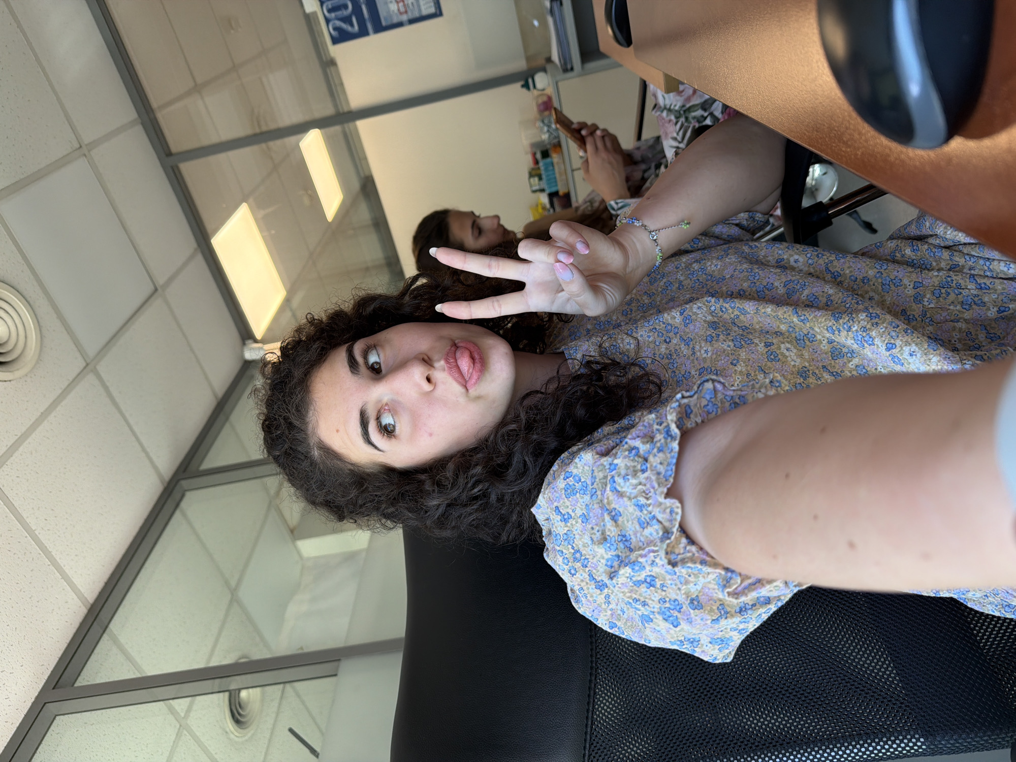 In Sofia, Bulgaria, an office environment is depicted. The foreground features a young woman with curly dark hair, wearing a floral print dress. She is seated in a black office chair and is making a peace sign with her right hand, her tongue sticking out. Her gaze is directed upwards and to the left. In the background, another young woman is visible, seated and looking at her phone. The office space is characterized by glass partitions, suspended ceiling tiles, and fluorescent lighting. A desk with a computer mouse is present in the immediate foreground.