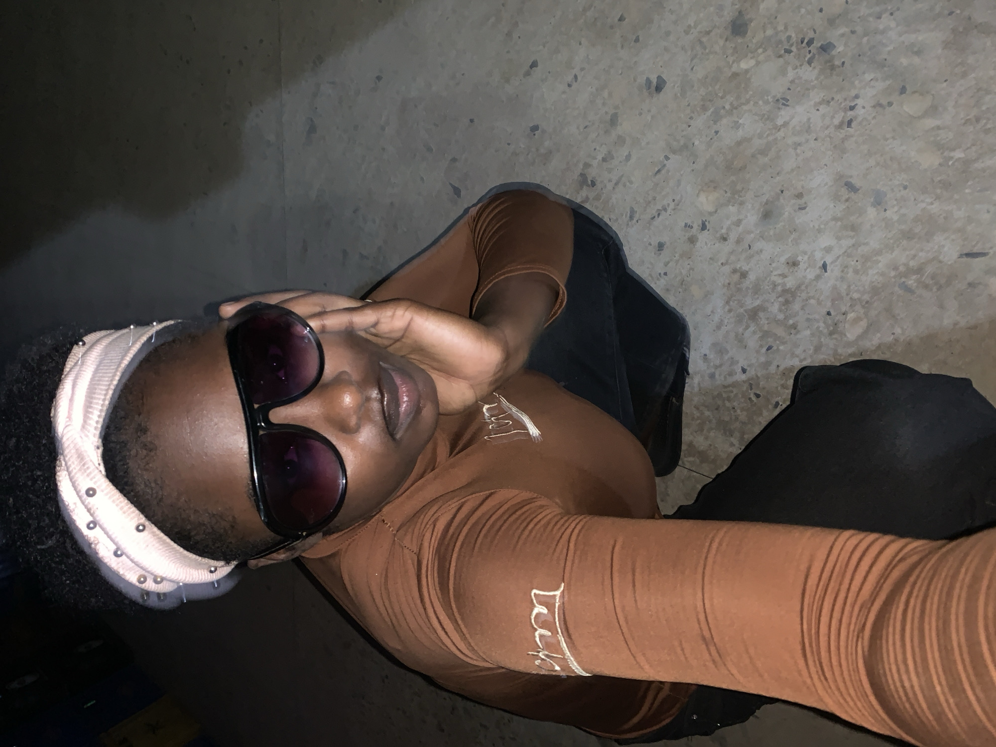 The image shows a person with dark skin and short, curly hair, wearing a pink headband and large, dark sunglasses. They are wearing a long-sleeved brown shirt with white lettering on the sleeve and chest. The person is wearing black pants. They appear to be taking a selfie, as their arm is extended towards the camera. The background is a textured gray surface and a dark, shadowed area. There is not enough information to confirm this location is New Stadium, Nigeria.