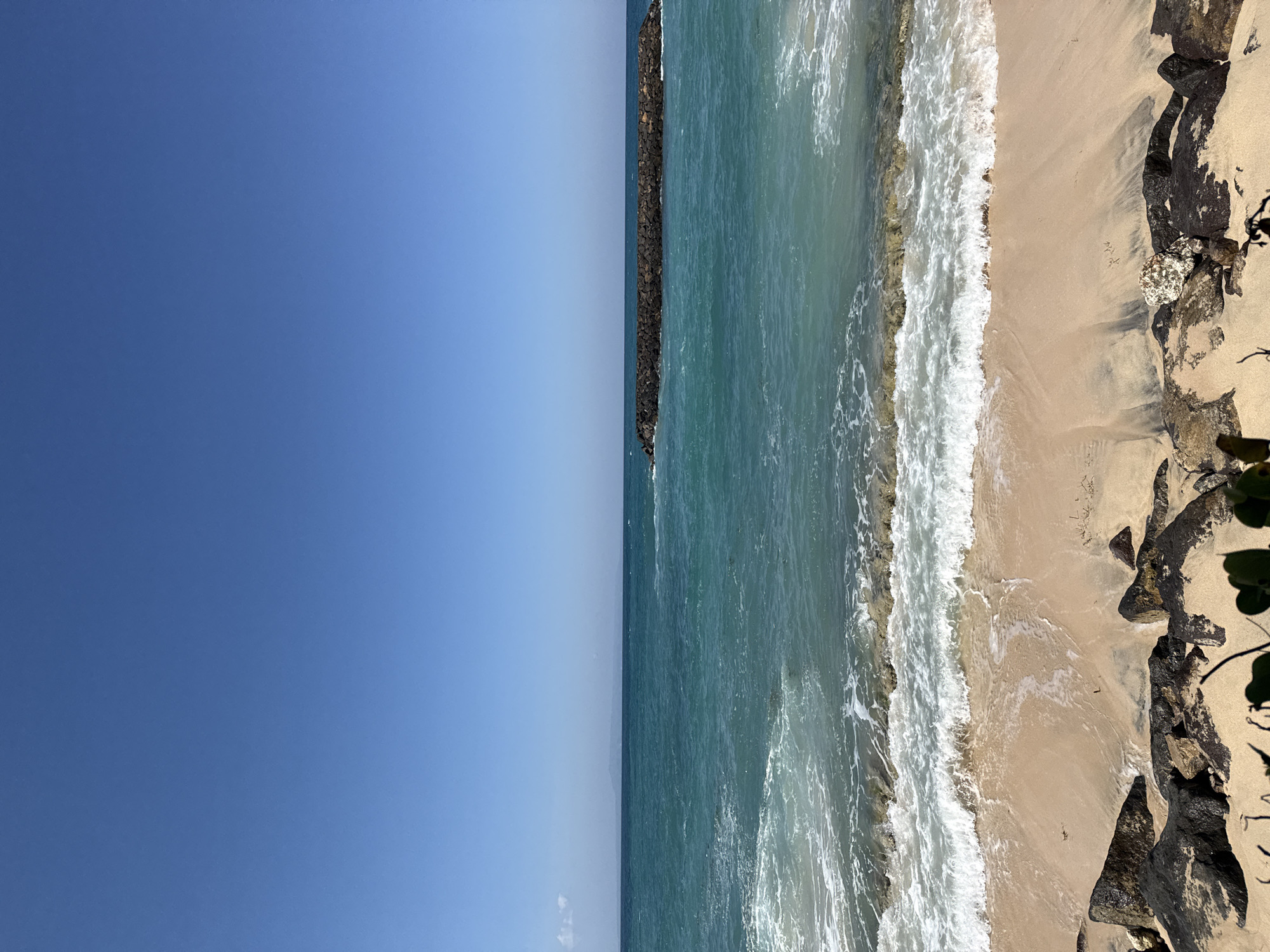 The image displays a coastal scene in Kuta Selatan, Indonesia, characterized by a clear, bright blue sky covering the upper half of the frame. The sky meets a distinct horizon line with the ocean, which transitions from a light turquoise near the shore to a deeper blue-green further out. Faint, distant land or mountains are visible on the horizon towards the left side. A dark, elongated rock structure, functioning as a breakwater or groyne, is situated horizontally in the midground of the ocean. White-capped waves are actively breaking onto a light tan sandy beach in the foreground. The immediate shoreline features scattered dark, wet rocks and patches of darker, damp sand. Minimal green foliage is present at the very bottom edge of the frame. No people or artificial structures, other than the breakwater, are visible. The lighting conditions indicate bright daylight.