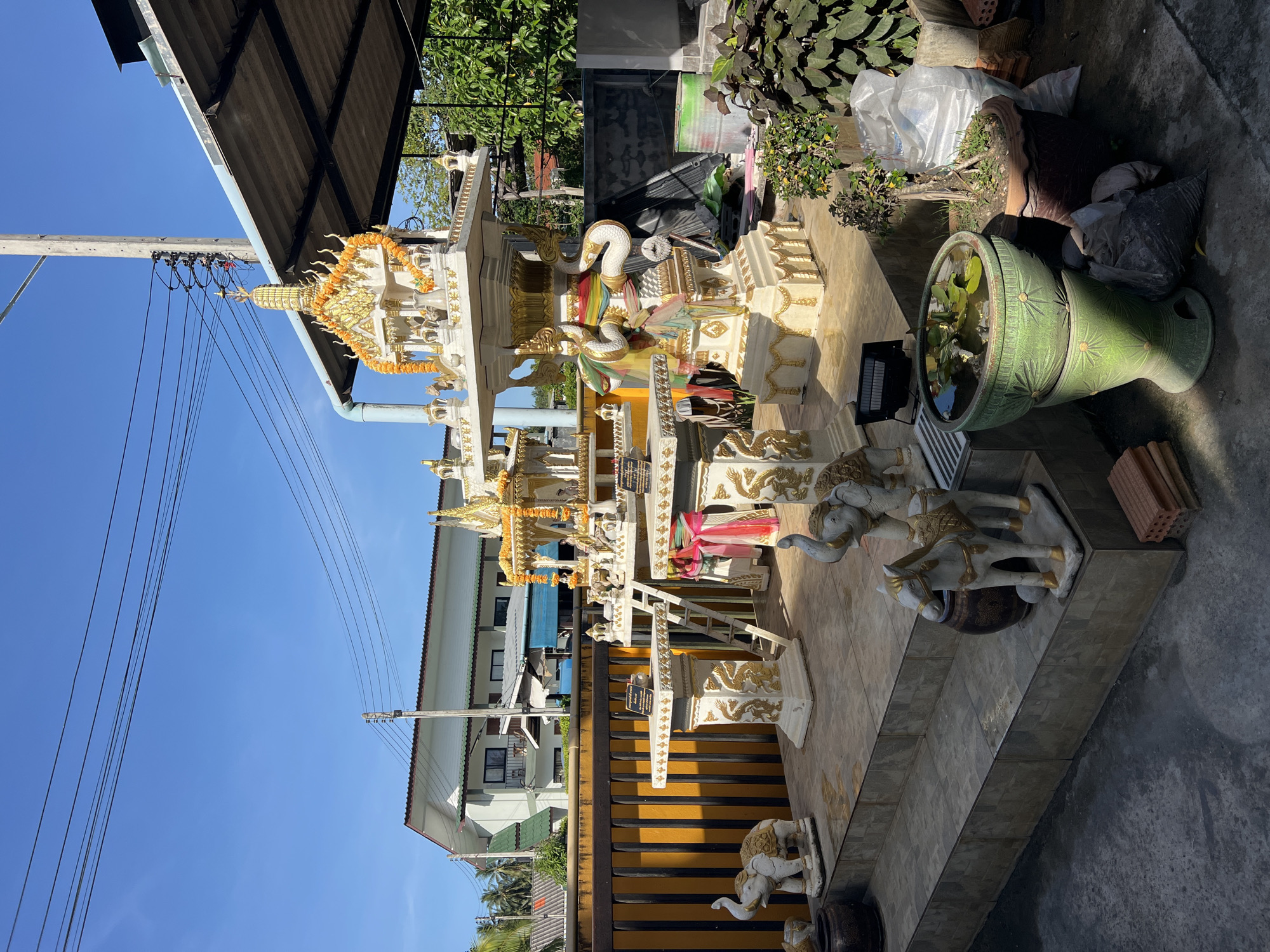 The image depicts a spirit house compound in Baan Lipa Noi, Thailand. The multi-tiered white and gold spirit house is adorned with garlands and snake figures. An assortment of statues, including elephants and horses, are positioned near the spirit house. Potted plants and miscellaneous items, such as bricks and bags, are also present. A building with a yellow staircase and an adjacent residential structure are visible in the background, against a clear blue sky. Power lines traverse the upper left portion of the frame.