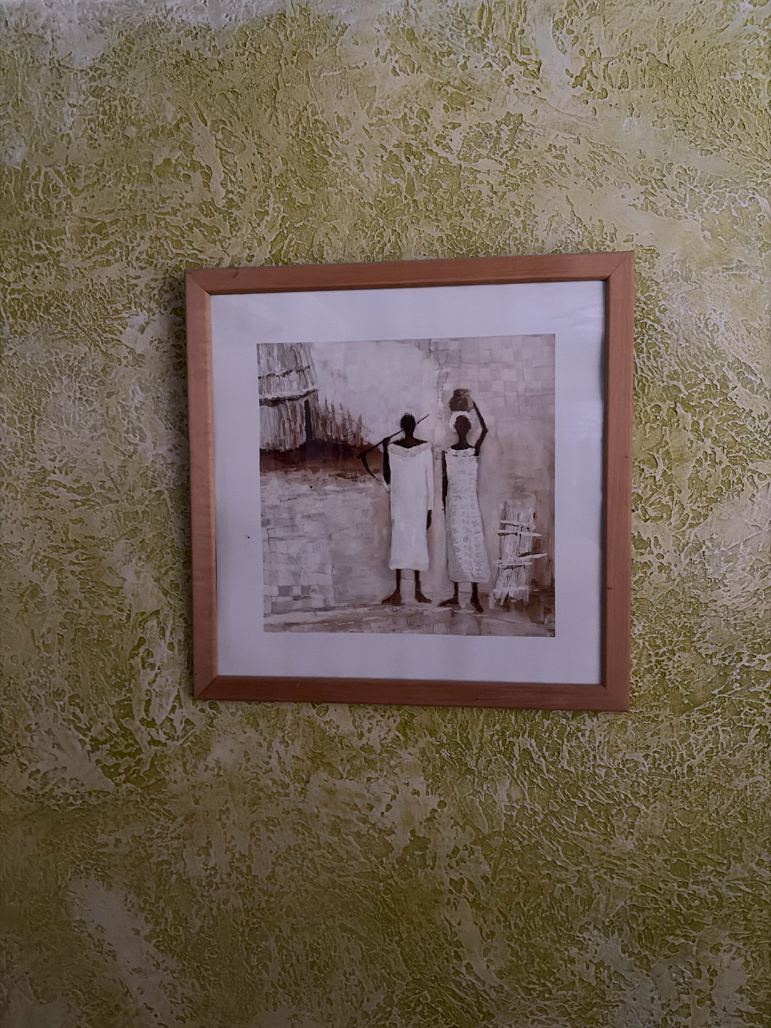 A framed painting hangs on a textured wall. The painting depicts two silhouetted figures, presumably women, in white garments against a muted background. One figure on the left holds a long object over their shoulder, while the figure on the right carries a pot on their head. To the right of the figures, a rustic wooden structure resembling a fence or drying rack is visible. The wall behind the painting has a mottled green and beige texture, creating a rough, uneven surface. The painting is housed in a simple wooden frame. The scene appears to be an interior shot, possibly in a home or gallery, located within the Livadiyske settlement council, Ukraine.