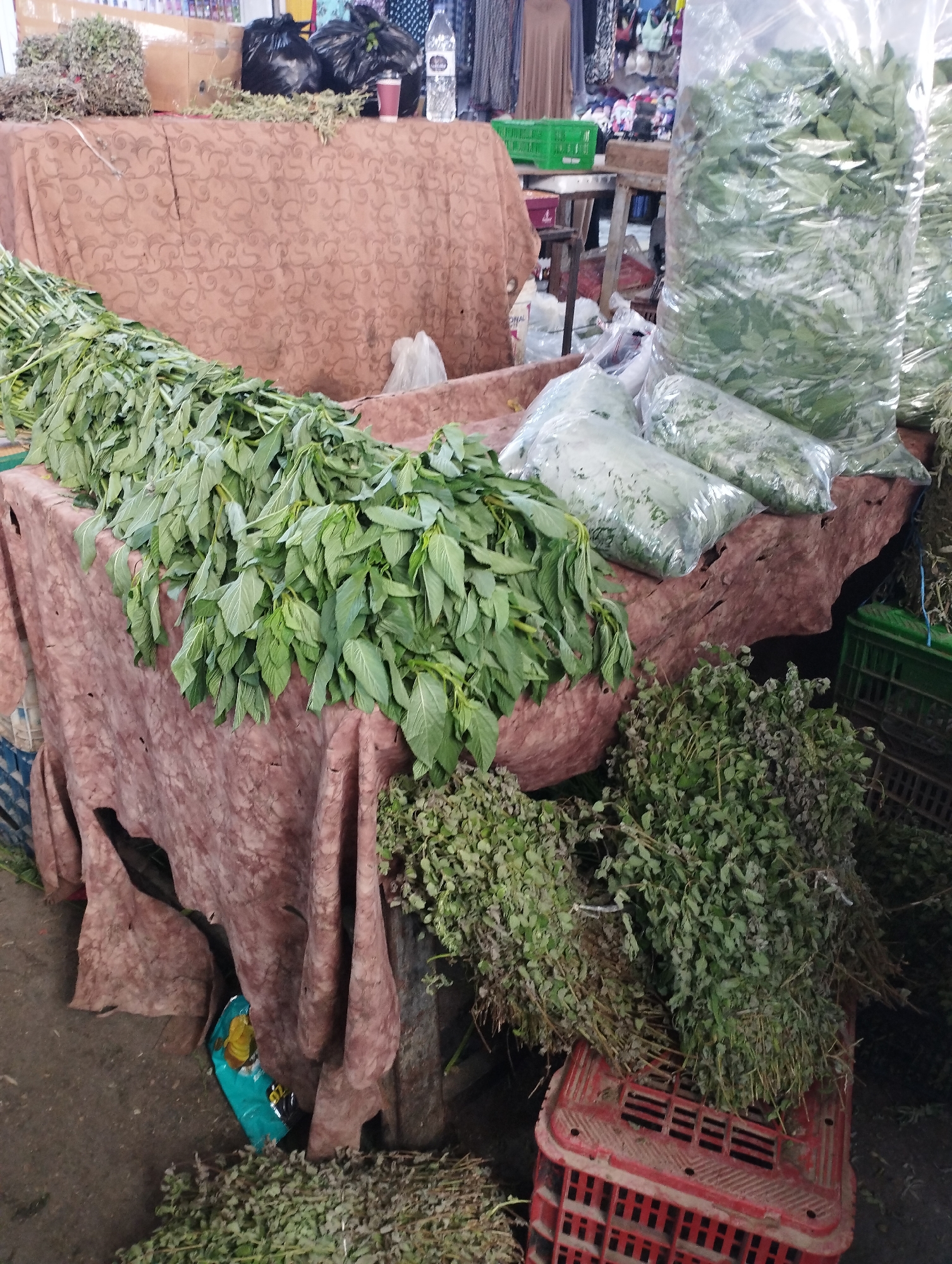 This image depicts an outdoor market stall in Baqa'a camp, Jordan. The stall is covered by a draped, patterned fabric and displays a variety of fresh produce, primarily herbs and leafy greens. In the foreground, a large pile of long, leafy green stalks is laid out on the fabric-covered table. To the right, several bundles of dried herbs, likely oregano or thyme, are arranged, some on a red plastic crate. In the background, large clear plastic bags filled with more green produce are visible, as are several green plastic crates and black trash bags. The ground is a dirt or concrete surface. The overall impression is of a vibrant, bustling marketplace.