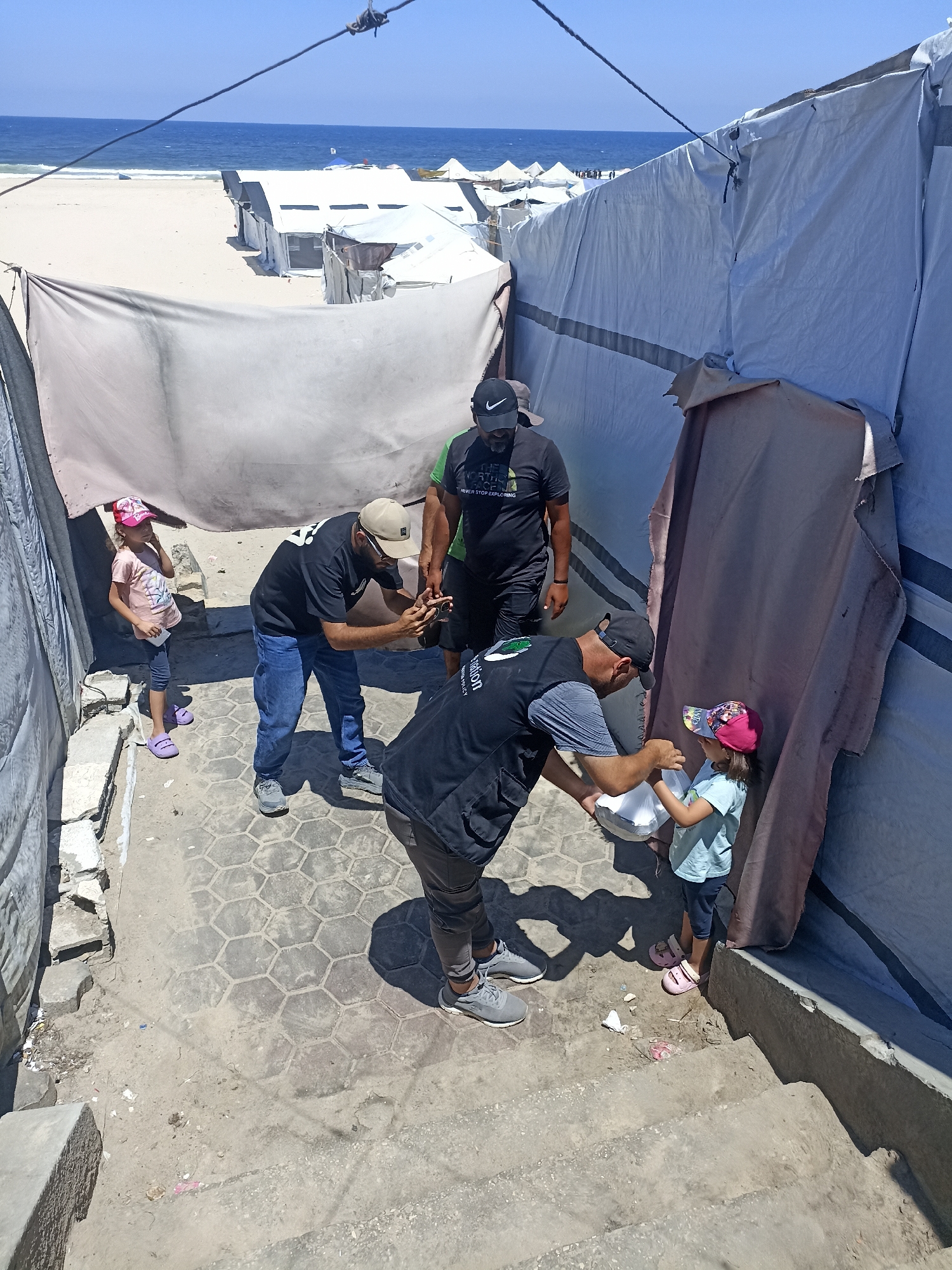 A group of people are gathered outside makeshift tents, likely in a refugee or displacement camp, near a beach. The scene is illuminated by bright sunlight, suggesting it's midday on a clear, sunny day. The ocean is visible in the background, with waves gently rolling onto the sandy shore. Several white tents with dark trim are set up on the beach, indicating a temporary living situation.
In the foreground, two men appear to be distributing items, possibly food or supplies, to two young children. One man, wearing a black t-shirt with a graphic and a beige cap, is holding a mobile phone and looking at it. Another man, in a black vest with a logo and a cap, is handing a white container to a young girl wearing a pink and blue cap and a light blue t-shirt. A younger girl, in a pink cap and a pink t-shirt, stands nearby. The ground is paved with hexagonal tiles, and there are some concrete steps and debris scattered around. The overall atmosphere seems somewhat somber, given the context of displacement, but the interaction with the children suggests an effort to provide aid. The vests worn by some of the men have text on them, but it is not clearly legible. The presence of tents near the sea implies a coastal location, and the general appearance of the setting aligns with images often associated with refugee camps in areas like Gaza.