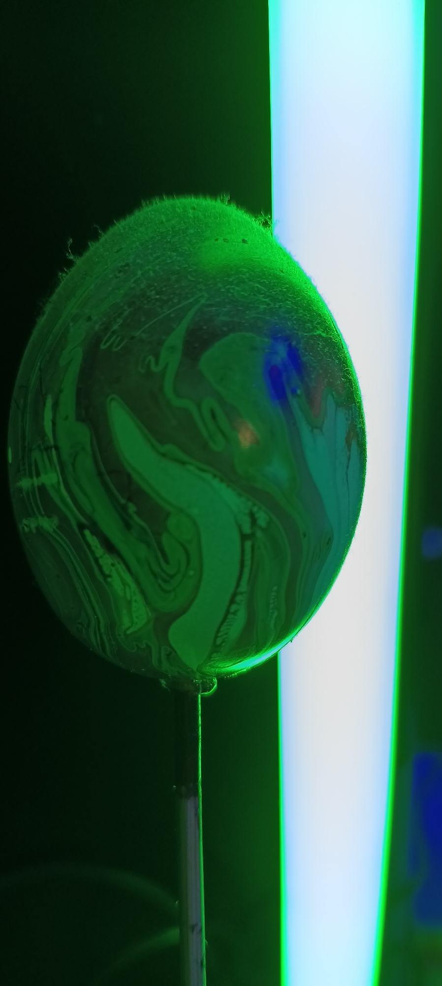 The image shows a close-up, macro photograph of a soap bubble suspended on a thin rod. The bubble is illuminated with green and blue light, creating iridescent patterns on its surface. The background is dark, with a bright, vertical light source to the right of the bubble. The bubble appears to be in the process of forming or dissipating, as evidenced by a small droplet hanging from its bottom. The image is characterized by its abstract and visually striking nature, focusing on the play of light and color on the thin film of the soap bubble. There are no people or identifiable landmarks in the image, nor any contextual information that would allow for localization within Wels, Austria.