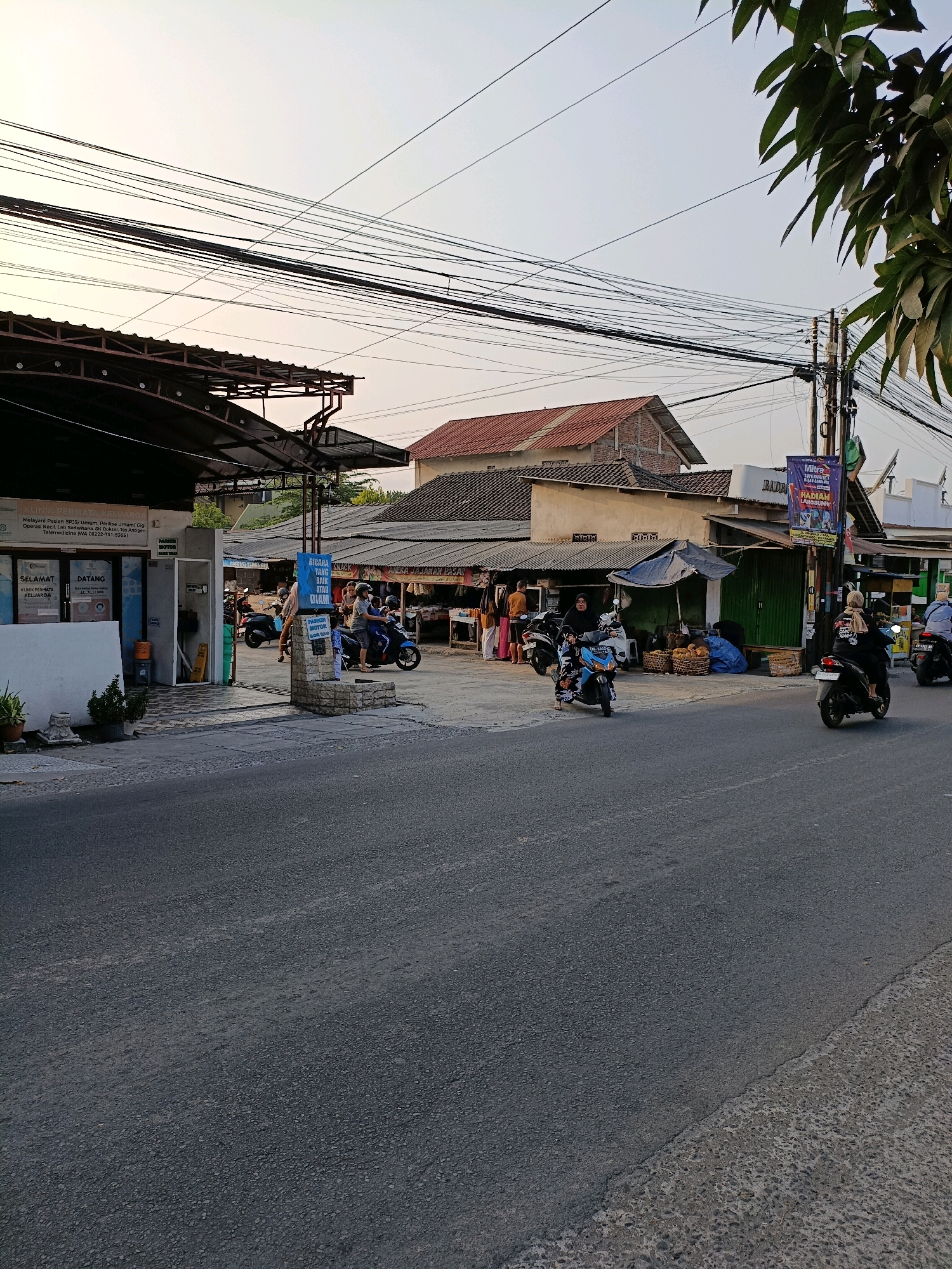 This image depicts a street scene in Purwomartani, Indonesia, likely during the late afternoon or early evening, given the lighting conditions. The foreground is dominated by a paved road. To the right of the road, a narrow sidewalk is visible, transitioning into a textured embankment. On the left side of the road, a building with a covered outdoor seating area or awning is present. This establishment appears to be a clinic, indicated by signage that includes "KLINIK PERMATA KELUARGA" (Family Gem Clinic) and text related to patient services. In front of the clinic, two potted plants are situated.
Beyond the clinic, a row of shops and stalls lines the street. Several individuals are seen interacting with these establishments. Motorbikes are a prominent mode of transport, with several parked and some in motion. Two motorbikes are parked in front of the clinic, and a blue motorbike with two riders is in the process of turning. Further down the street, a dark-colored motorbike with a single rider is moving away from the camera, and another dark motorbike with a rider is parked. Several people are walking and standing near the shops.
The shops themselves feature open fronts, displaying goods such as food and other items in baskets. One stall has a blue awning. A tall pole with multiple electrical wires running across the upper portion of the image is a notable feature of the urban landscape. The sky above is a pale, hazy blue. A banner with text and an image is hanging from the pole in front of one of the stalls. Green foliage from a tree is visible in the upper right corner of the frame. The overall impression is of a busy, functional commercial area within the town.
