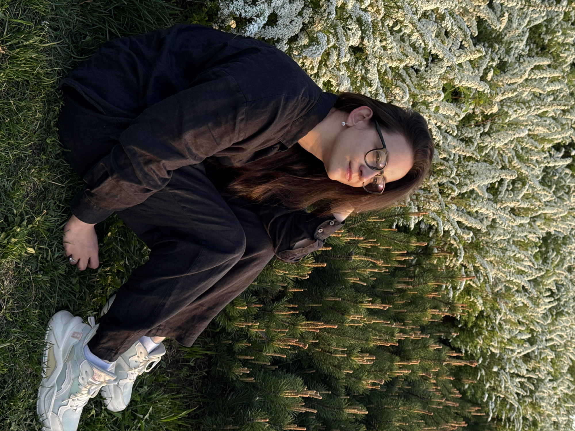 A young woman with long brown hair and glasses is sitting on grass next to a bush. She is wearing a black button-down shirt and black jeans, and white sneakers with light blue accents. She is looking towards the camera with a relaxed expression. The background includes green grass, a pine bush, and a bush with many small white flowers. Based on visual clues and the prompt, it is possible this photo was taken in or near Nizhny Novgorod, Russia, in a natural or park-like setting. The lighting is bright, suggesting the photo was taken outdoors during the day.