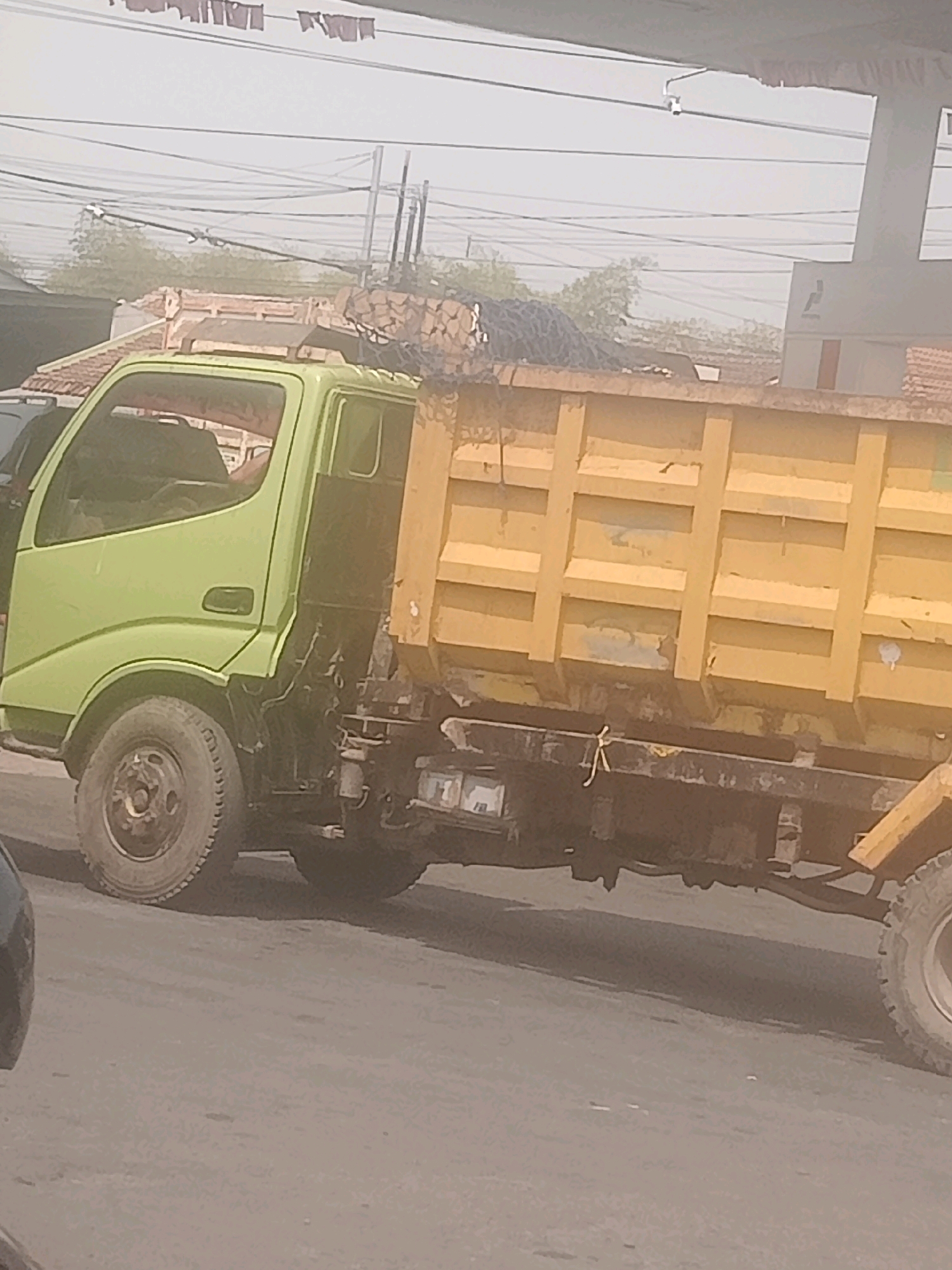 A light green dump truck is parked on a paved road in Lebaksiu, Indonesia. The truck has a yellow dump bed that appears to be filled with dark material, possibly coal or gravel, and is covered with a net. The truck is positioned at an angle, with its front on the left and its rear on the right. The cab of the truck is visible, and appears to be empty. Behind the truck, there is a low building with a red tile roof, and beyond that, trees and utility poles with overhead wires. The sky is overcast and hazy. The overall scene suggests an industrial or commercial area.