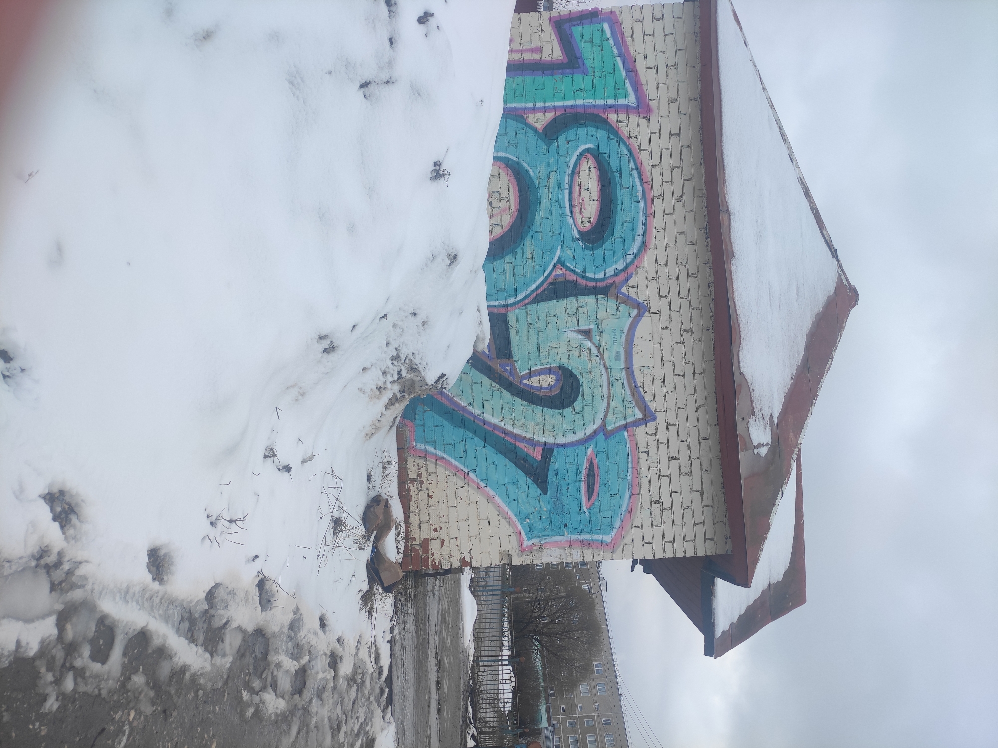 A vertical, eye-level shot depicts a wall with graffiti, partially obscured by snow, located in Chaikovsky, Russia. The graffiti, rendered in shades of blue, turquoise, and magenta, appears on a light-colored brick wall, likely an exterior surface. A substantial snowdrift rests against the left side of the wall, obscuring the lower portion of the graffiti. The top of the wall extends into a roof overhang, also covered with snow.
The foreground features a snow-covered ground with visible footprints. In the background, elements of a city landscape can be observed, including buildings and trees, suggesting an urban setting. The sky is overcast. No people are visible in the image.