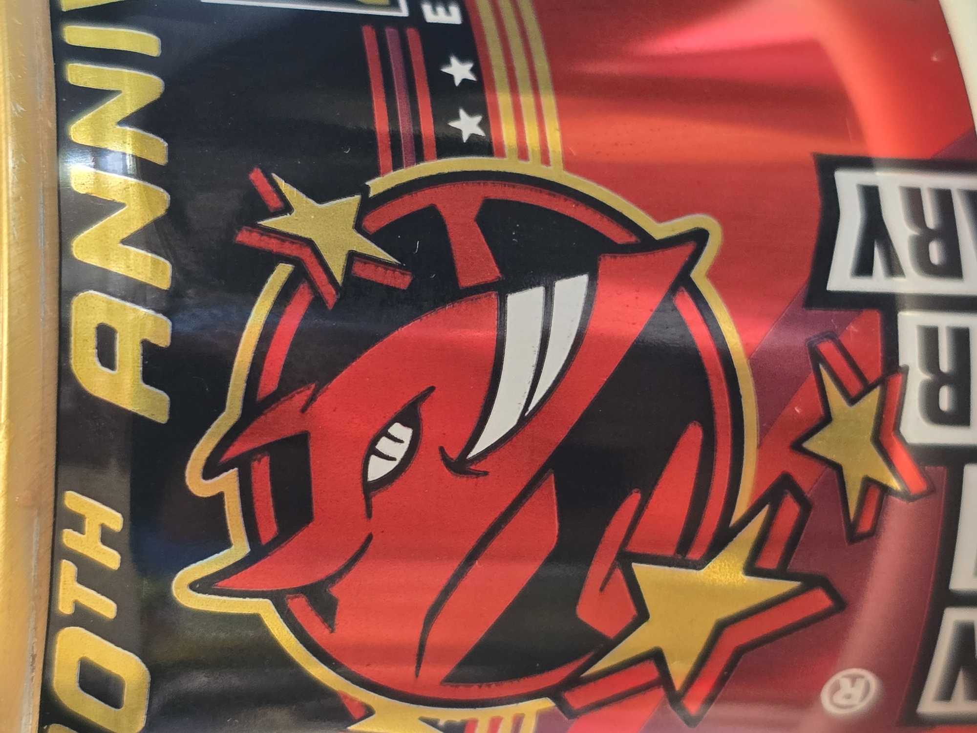 A close-up shot captures a vibrant, glossy design, likely from product packaging or a label, featuring a dominant red, black, and gold color scheme. The central element is a stylized red devil or demon head logo, outlined in gold, with white teeth and a single visible eye, conveying a fierce or mischievous expression. This logo is set within a black circular area.
Surrounding the central emblem are several prominent gold stars, some accentuated with red outlines, contributing to a celebratory aesthetic. Arcing red and gold stripes appear in the upper portion of the design, resembling a banner or decorative element. The surface exhibits a metallic sheen, reflecting light, particularly across the red areas.
Partially visible text in gold, oriented vertically along the left edge, reads "TH ANNIVE", strongly suggesting the word "ANNIVERSARY". On the right side, white letters with a black outline, also vertically oriented, show "RY", further confirming "ANNIVERSARY" as part of the overall text. A registered trademark symbol (®) is faintly visible near the bottom right. The design appears to be commemorating an anniversary.