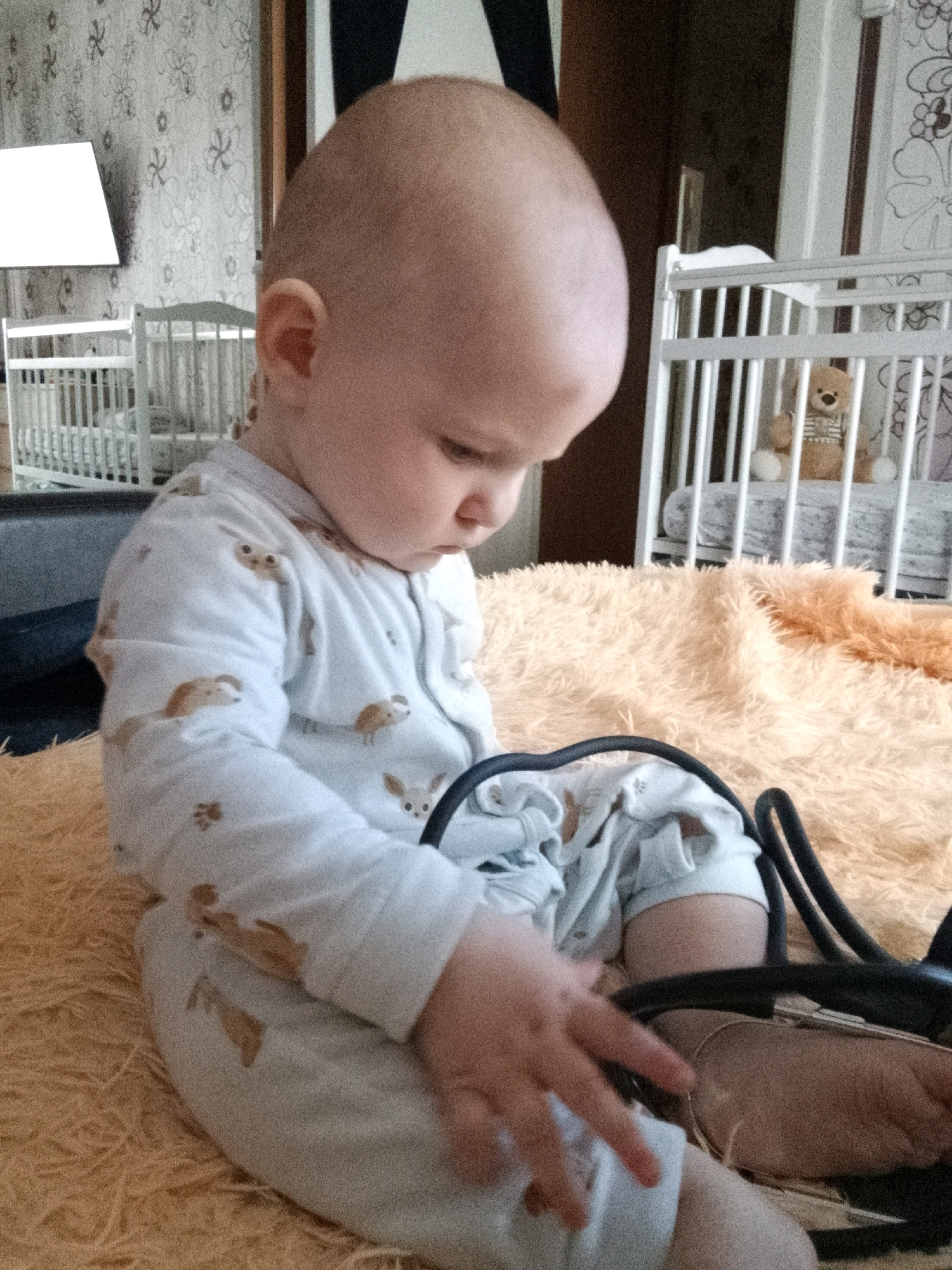The image is a close-up shot of a bald baby sitting on a fuzzy, peach-colored blanket. The baby is wearing a white onesie with a pattern of brown cartoon dogs. The baby is looking down and to the right, with their left hand reaching towards their lap. A black object, possibly headphones or a toy, is partially visible in the foreground, obscuring the baby's legs. In the background, out of focus, are two white cribs and a portion of a patterned wallpaper. The lighting is dim, suggesting an indoor setting. There is no information to suggest the location is in Minsk, Belarus.