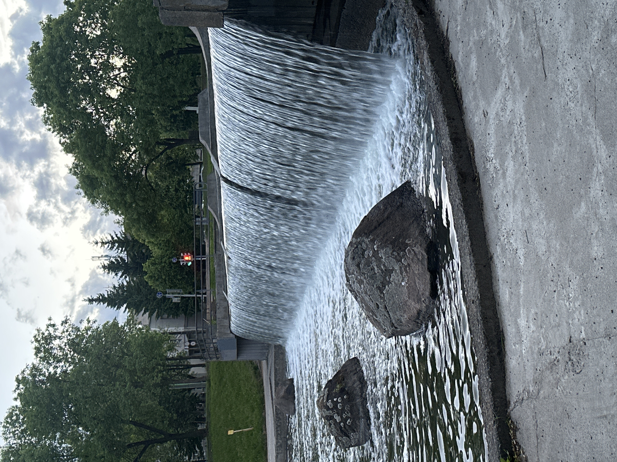 The image shows a waterfall structure in the city of Minsk, Belarus. The waterfall is constructed of concrete, with water flowing over a curved edge into a pool. Two large rocks are positioned in the pool. In the background, trees and a traffic light are visible. A pedestrian walkway and fence are adjacent to the structure. The sky is overcast.