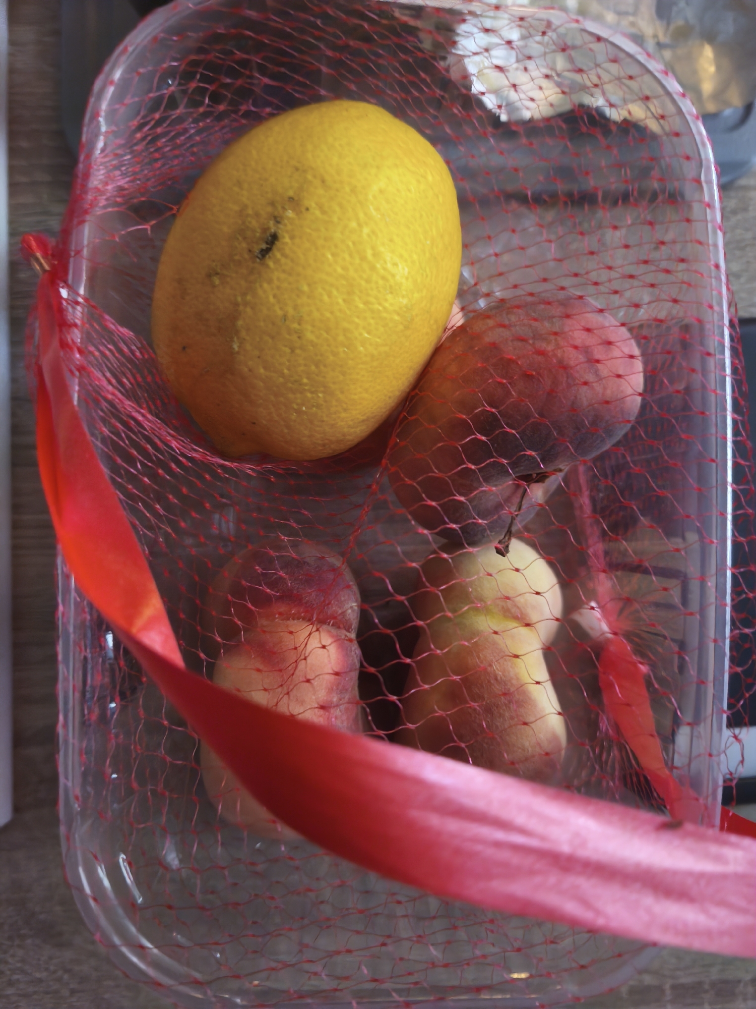 A clear plastic container holds a yellow lemon and two peaches, all encased in a red mesh bag tied with a red ribbon. The container is placed on a wooden surface. The scene is indoors, likely in Assen, Netherlands, suggesting a domestic setting such as a kitchen counter.