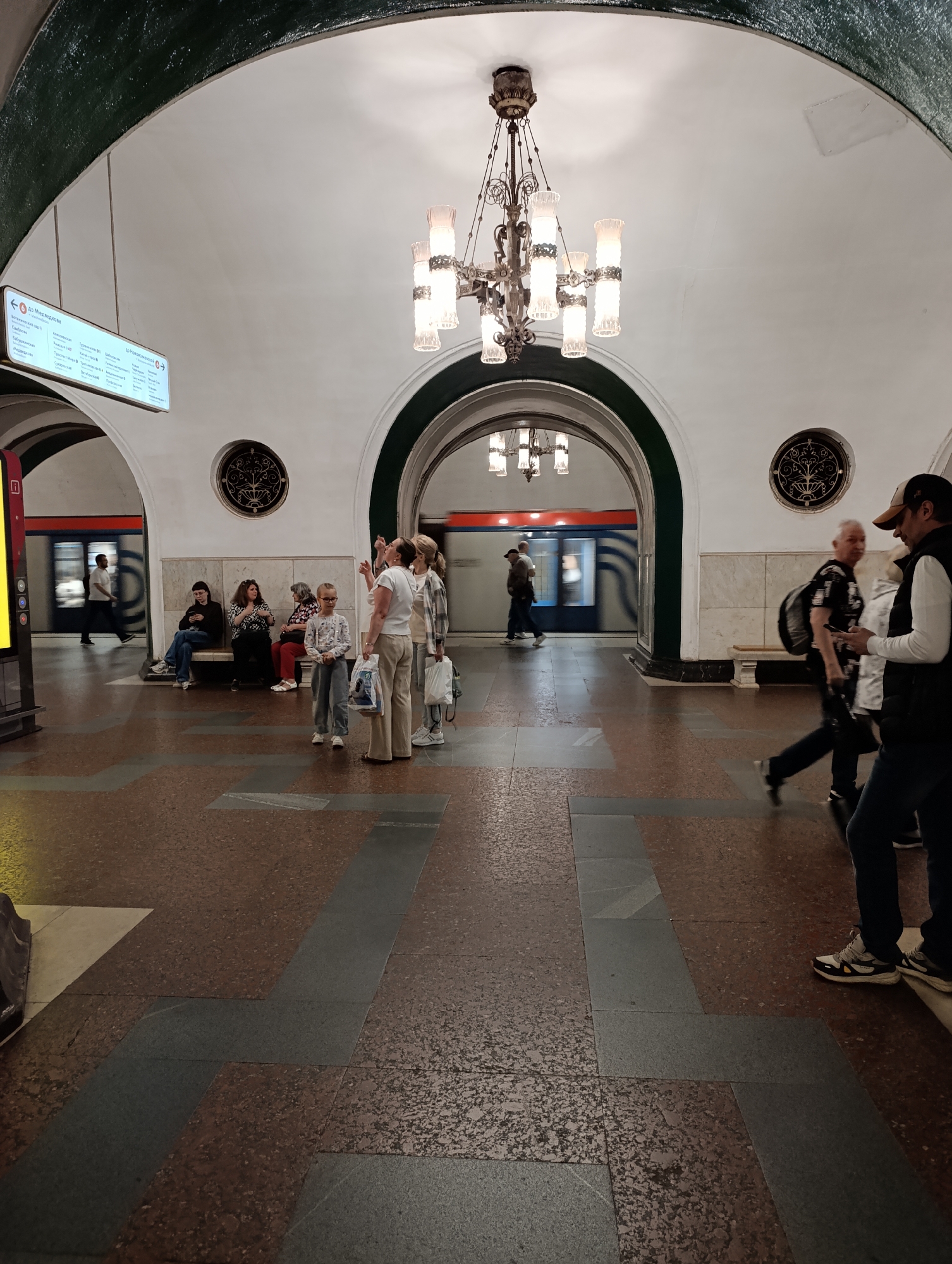 The image depicts a grand, ornate subway station, characteristic of the Moscow Metro in Russia. The scene is dominated by vaulted ceilings and large arched passages, illuminated by decorative, multi-bulb chandeliers hanging from above. Another similar chandelier is visible through the central archway, which leads to the train platform.
Numerous people are present in the station. In the middle ground, a group of women and a young girl stand near the central archway, while another group of women and a girl are seated on a wooden bench to the right. One woman looks upwards. In the foreground on the right, two men are walking, one wearing a baseball cap and dark vest, the other a patterned shirt and backpack; both show signs of motion blur. On the platform beyond the central arch, a blue and red subway train is in motion, also blurred, with two male figures visible nearby. Other people are seated on a bench to the left.
The station floor is covered in a distinctive pattern of reddish-brown and dark grey granite or stone tiles. Circular, ornate decorative elements are embedded in the walls on either side of the central archway. The walls are light-colored, with dark green accents along the lower edges of the arches.
On the left wall, a bright digital sign displays Cyrillic text. The most prominent line clearly indicates "← до Медведкова" (to Medvedkovo), along with a list of other station names below it, functioning as a directional guide for passengers. The artificial lighting suggests an indoor setting, likely during operational hours. People appear engaged in typical commuting activities, either waiting or moving through the station.
