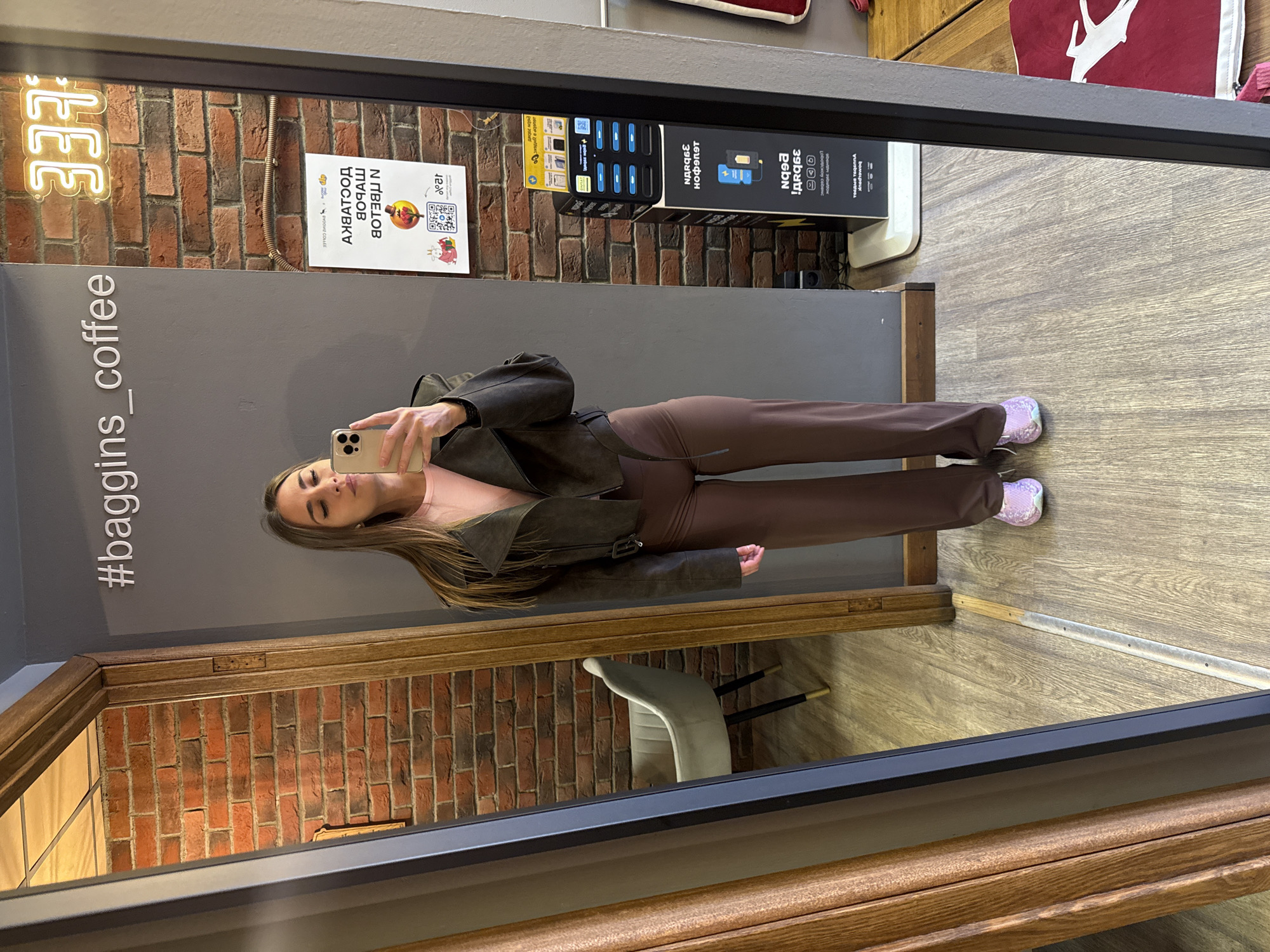 The image depicts a woman in a mirror selfie, seemingly within a cafe. The woman is wearing brown pants, a nude top, and a brown jacket. Her phone is in her hand, held up for the selfie. The cafe interior includes brick walls, a gray wall with the hashtag "#baggins_coffee," a neon sign reading "337," a promotional sign written in Russian with the words "Ν ΠΒΕLOB ПОЧАШ АХВАТООД" and the phrase "Lobotickoy cebenc," and a mobile charging station. The floor is wooden. The reflection in the mirror reveals a chair and additional parts of the cafe.
Based on the Russian text and the "#baggins_coffee" hashtag, it is reasonable to assume that the location is in Saint Petersburg, Russia.