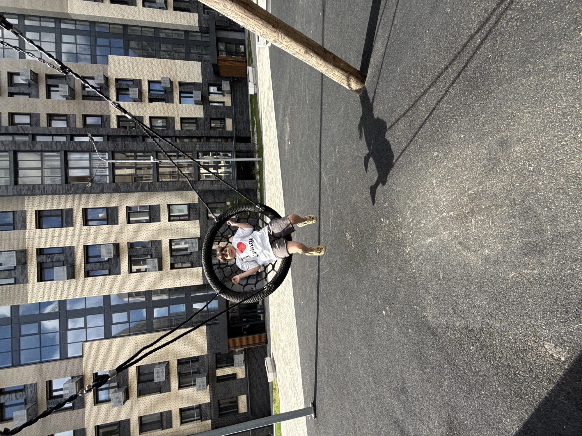 A child is reclining in a black net swing, suspended from a wooden pole in an urban playground. The ground around the pole and swing is paved with dark asphalt. The child wears a white t-shirt with a red heart design, gray shorts, and yellow shoes. The swing's shadow is visible on the asphalt. In the background, there is a multi-story building with a combination of tan brick and dark gray window frames. The location is a city of Unknown, Unknown.