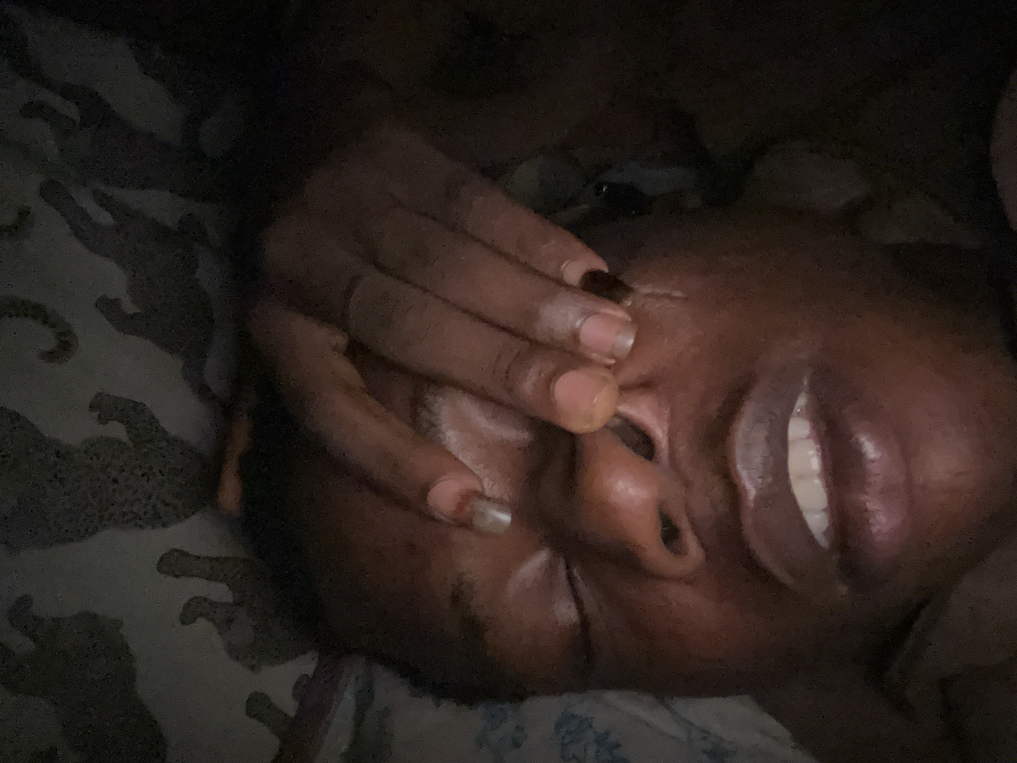 A close-up, low-angle shot depicts a person lying down with a hand covering their eyes. The person's skin tone is dark, and their lips are parted, revealing their teeth. The fingers of the hand covering their eyes have manicured nails, with some painted a light pink color and one painted black. The background shows a patterned surface, possibly bedding. The environment appears to be dimly lit. The location is unknown.
