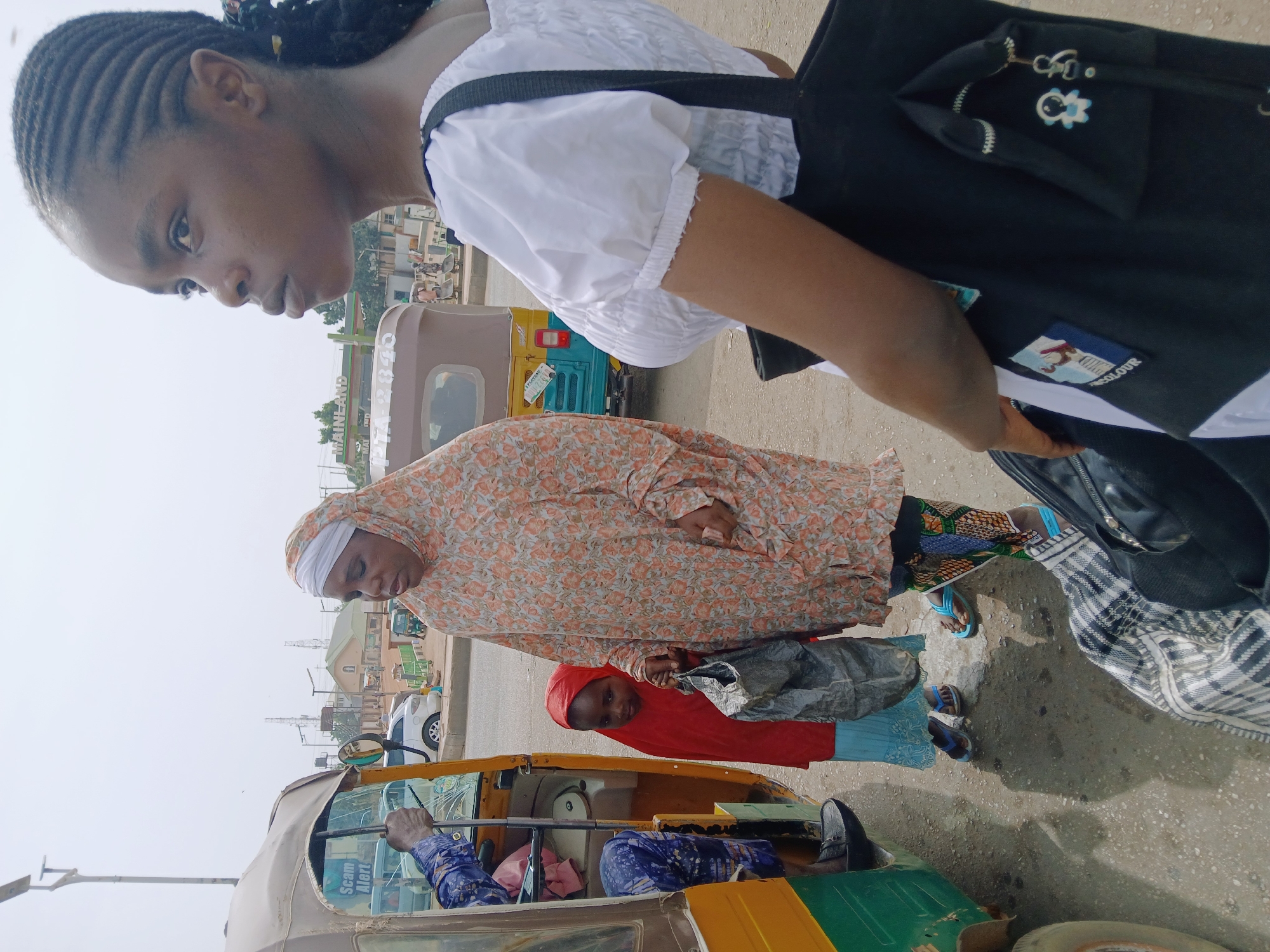 The image captures a street scene in Jos, Nigeria. A young woman in a white blouse and black shoulder bag is in the foreground. Behind her, a woman in a floral hijab and a child in a red hijab stand near a yellow and green auto-rickshaw, commonly known as a "keke." The auto-rickshaw driver is visible inside. Other auto-rickshaws and buildings are in the background. The sky is overcast.