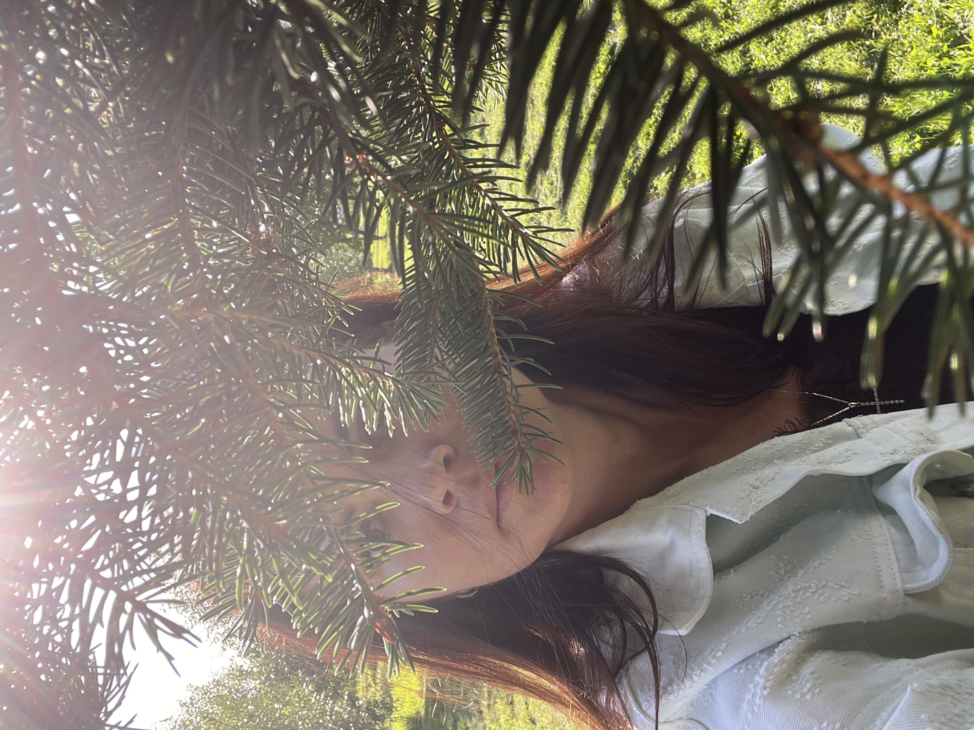 In this close-up, outdoor, daylight shot, a woman with auburn hair is partially obscured by the needles of a coniferous tree. She is wearing a light blue denim jacket over a black top with a delicate necklace. Her gaze is directed towards the camera, and her expression is neutral. The background features a sunlit grassy area, with hints of trees and foliage in the distance. The lighting suggests a sunny day, with the sun's rays creating lens flares. The setting appears to be a park or garden, possibly within the city of Saint Petersburg, Russia, given the overall atmosphere and the presence of manicured greenery.