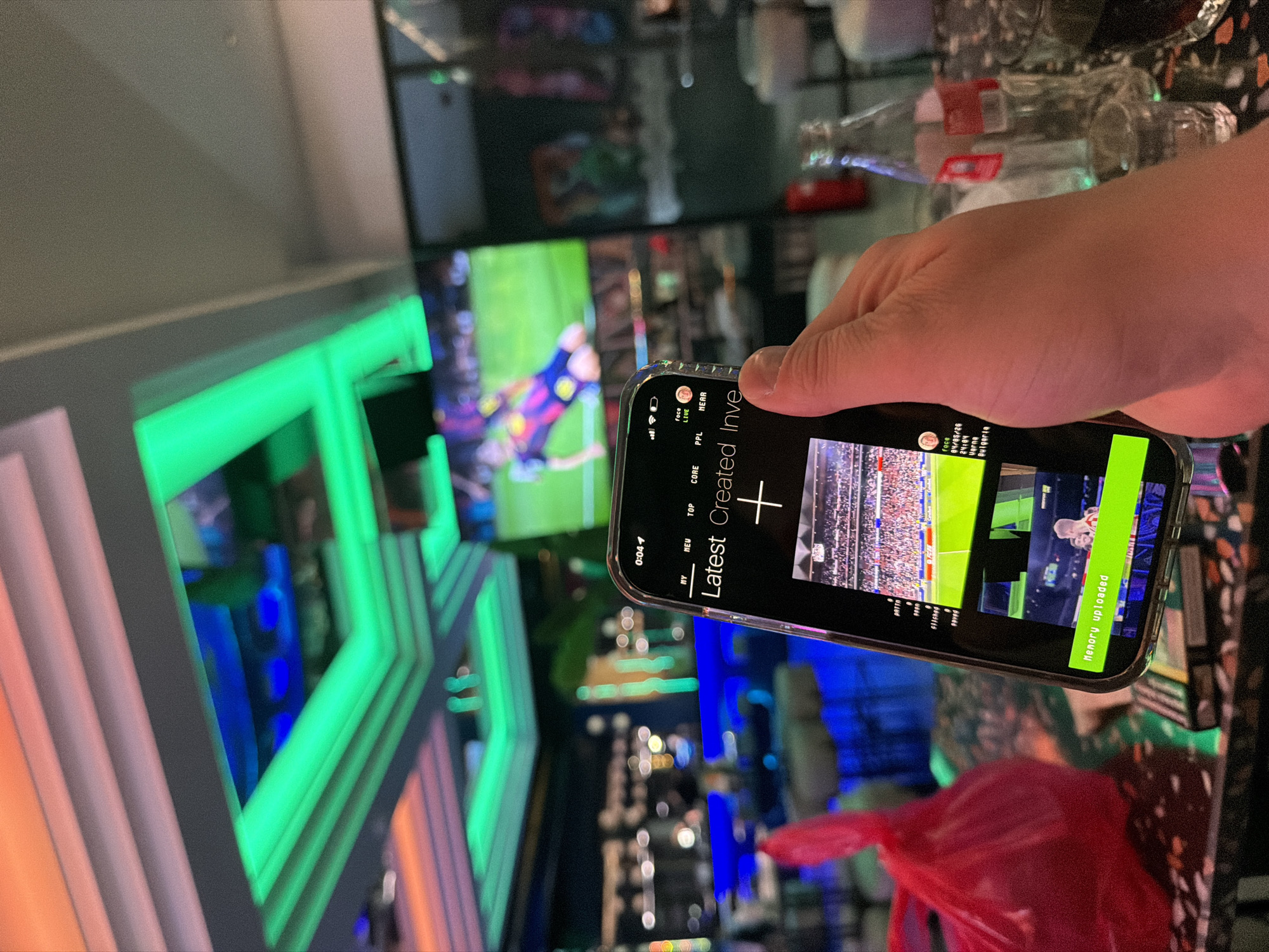 The image shows a person holding a smartphone in front of a bar setting. The smartphone displays a screen with the words "Latest Created Inve" and a "+ "sign. Below these words, there are several thumbnails of various videos, including one of a football match and another of a person. The phone also displays text mentioning "Varna" and "Bulgaria". In the background, there's a blurry view of a TV screen showing a football match, as well as neon lights, bottles, and decorative objects typical of a bar. The presence of "Varna" and "Bulgaria" on the smartphone screen suggests the location might be in Varna, Bulgaria.