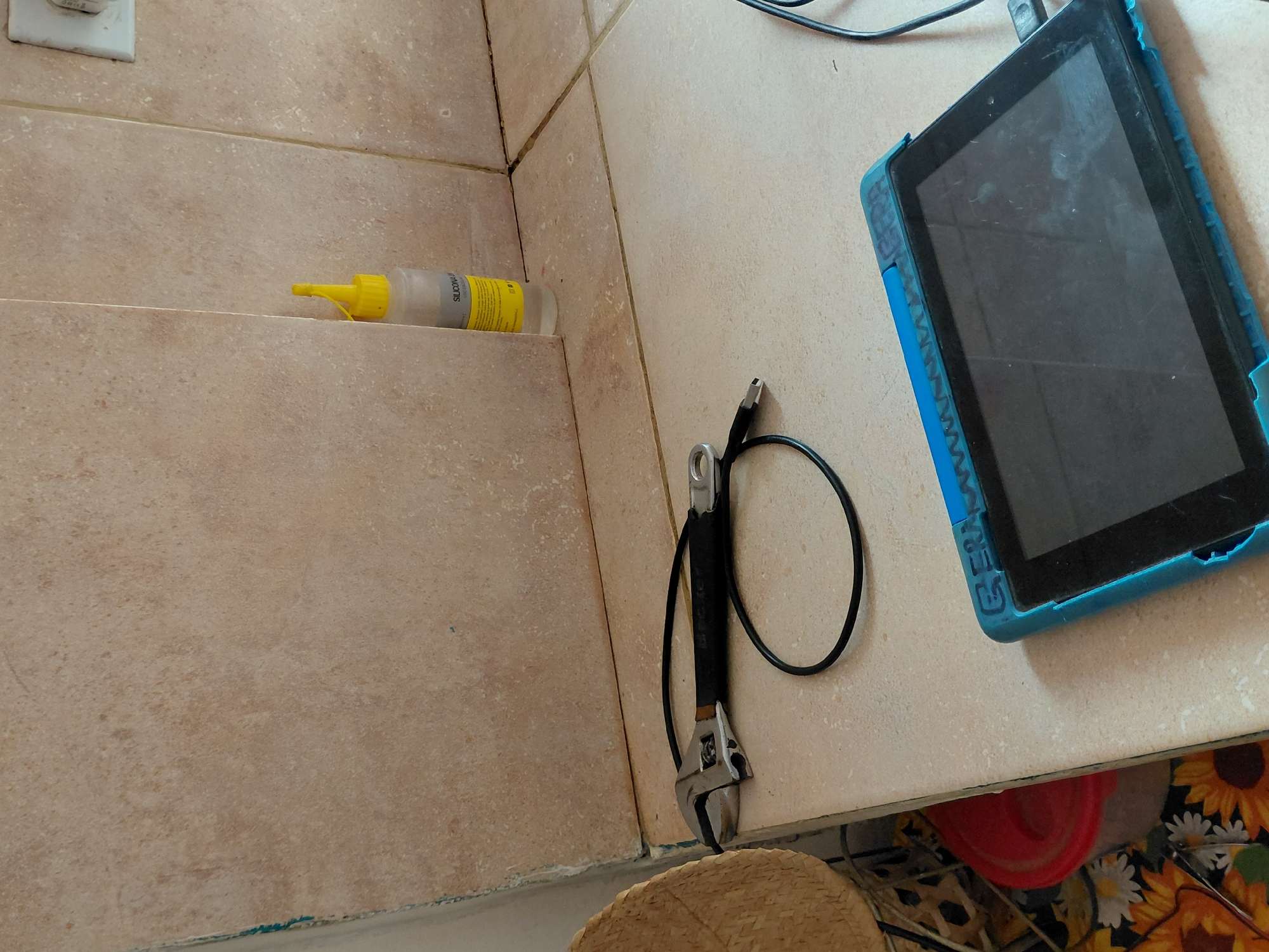 This image captures a still life scene in an indoor setting, likely a utility area or a space undergoing maintenance, located in Santa Ana, Venezuela. The primary subjects are various objects arranged on light brown or beige tiled surfaces.
In the upper right, a tablet encased in a vibrant blue, textured protective cover rests on a countertop. Its screen is dark and appears smudged or dusty. A black cable, possibly for charging, is plugged into the tablet. Adjacent to the tablet, lying on the tiled surface, is a silver and black adjustable wrench with a black cable looped around its handle.
To the left, on a lower tiled shelf or ledge, sits a clear plastic squeeze bottle with a yellow cap and nozzle. The bottle contains a clear liquid, and part of its yellow label is visible, displaying the text "SILICONA" (Spanish for silicone). An electrical outlet is mounted on the tiled wall in the upper left corner.
The tiles throughout the scene exhibit variations in tone and texture, suggesting a functional, rather than purely decorative, space. The grout lines, particularly in the corner where the wall and shelf meet, appear dark. Below the counter, peeking into the frame, are glimpses of a woven, straw-like object (possibly a hat or basket), a red plastic bucket or container, and a patterned fabric featuring sunflowers and daisies.
No human subjects are present, and no specific activity is taking place; the image presents an arrangement of tools and devices. The overall impression is that of a workspace or storage area where items are kept ready for use.
