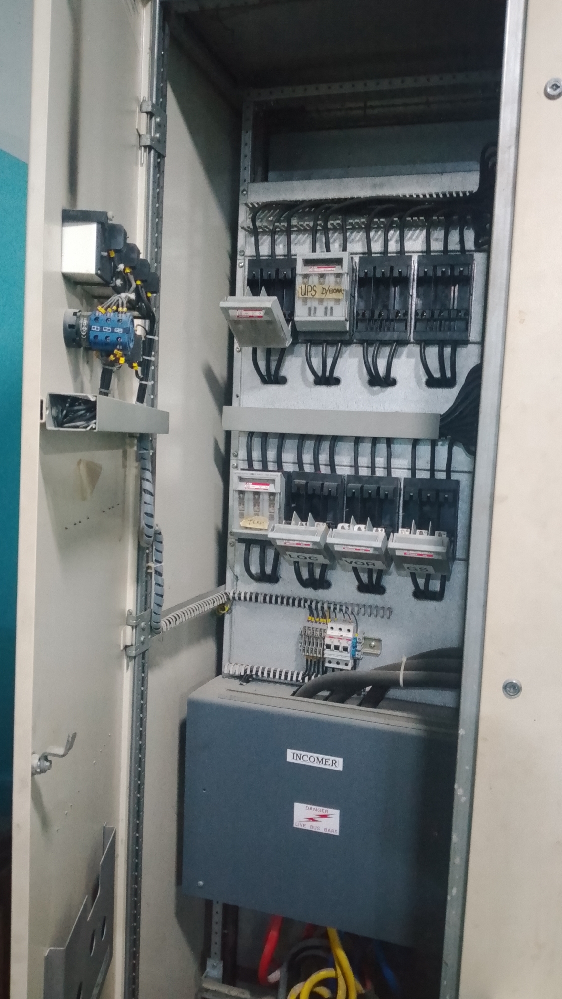The image shows an open electrical panel, likely located in Effurun, Nigeria based on the prompt. The panel is metallic and contains various electrical components including circuit breakers, fuses, wiring, and a device labeled "INCOMER" with a "DANGER LIVE BUS BARS" warning sticker. The wiring is a mix of black and yellow cables. A blue rotary switch with associated wiring is mounted to the left side of the panel. The overall environment appears industrial. No people are visible in the image.
